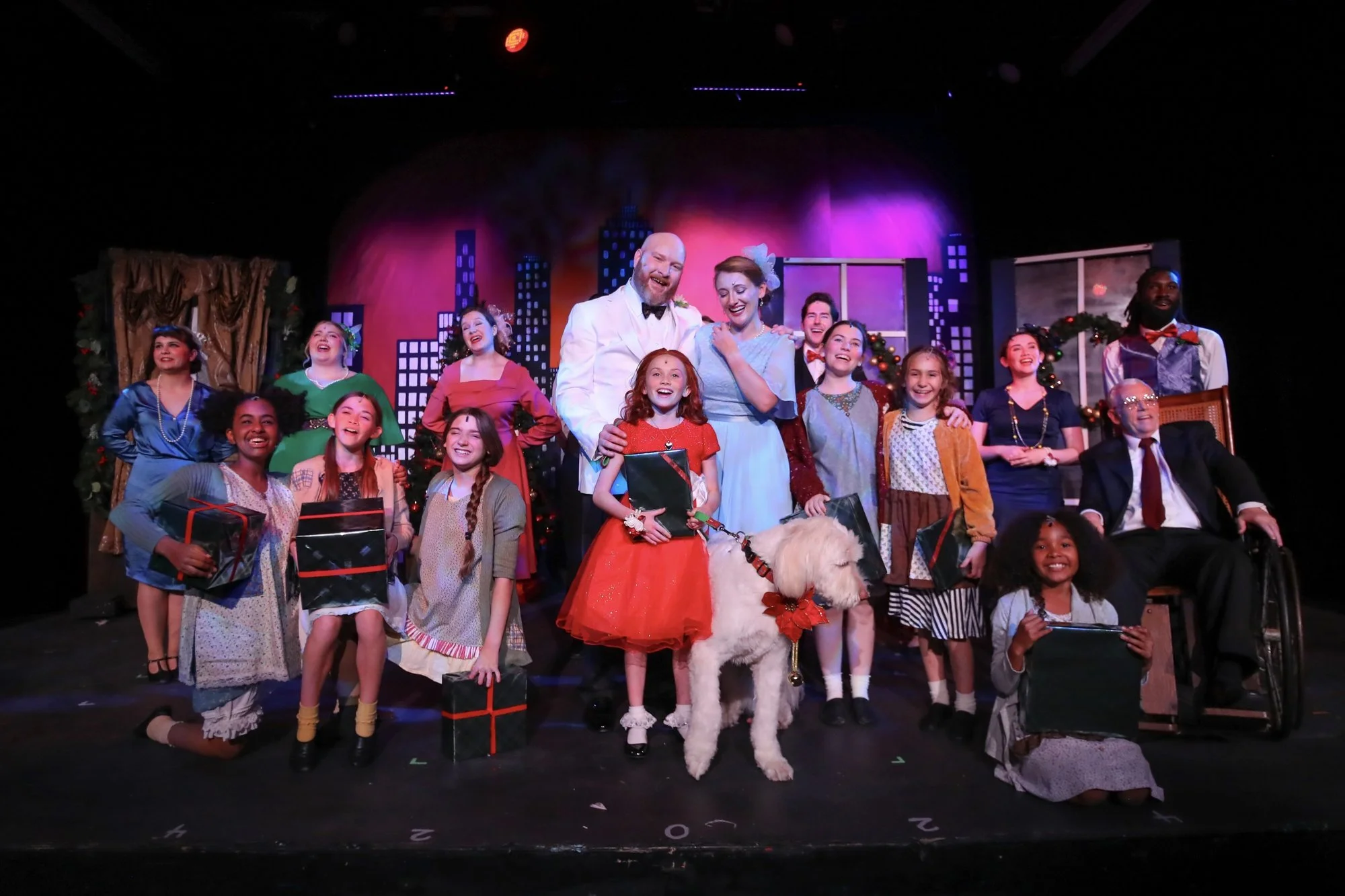 Stage with group of performers and children, festive holiday setting, skyline backdrop, holiday decorations, and a large white dog in the center, celebrating after a performance.