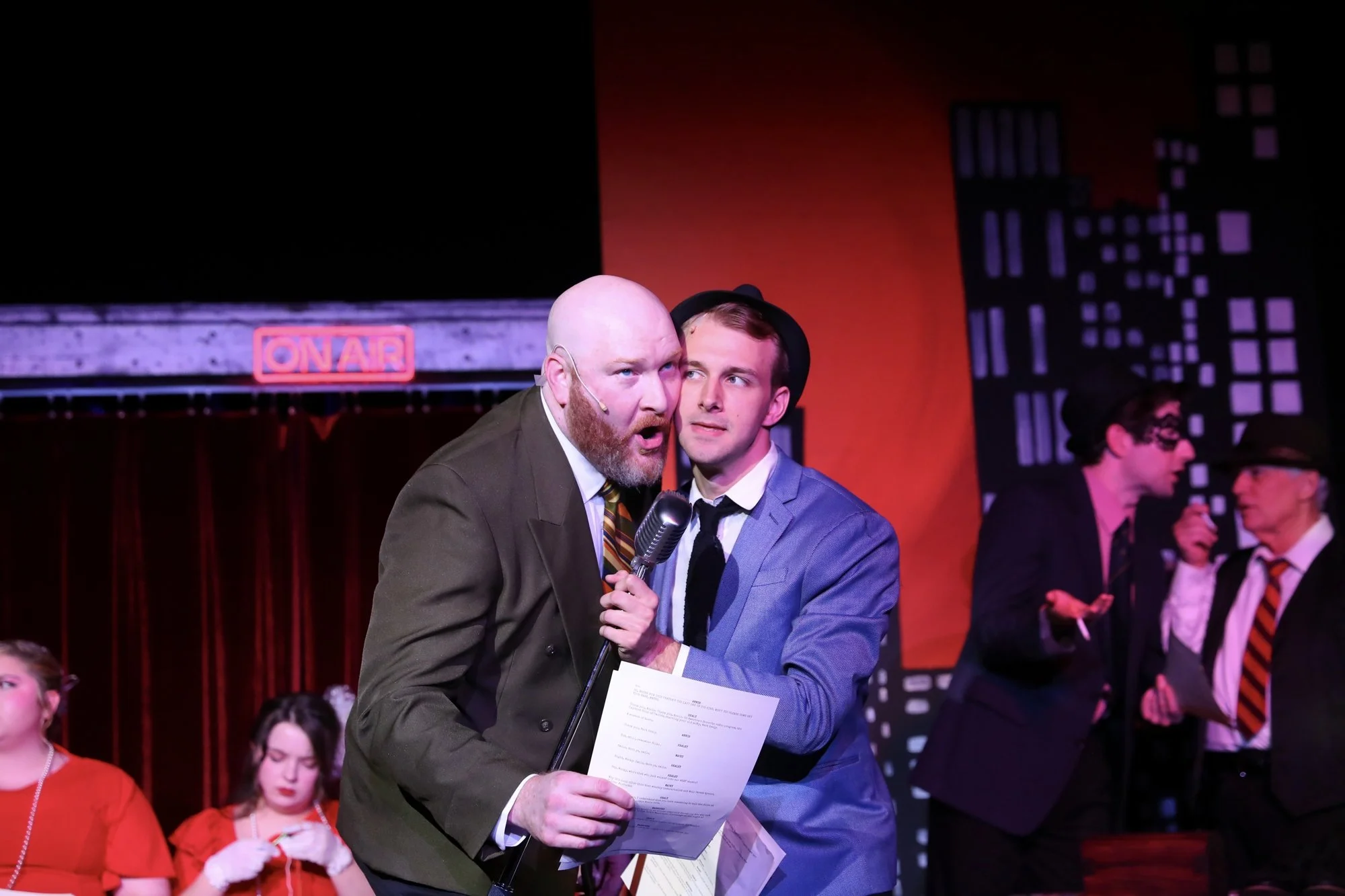 Two men on stage, one with a bald head and beard speaking into a microphone, writing on a paper, another man in a blue suit and black hat looking at him, with a red cityscape backdrop and an 'ON AIR' sign.