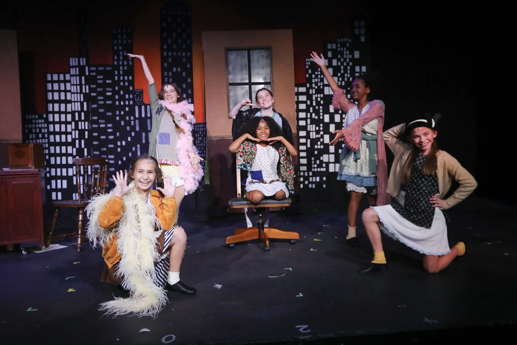 Group of young girls performing on stage in costumes, with a cityscape backdrop painted on the wall behind them.
