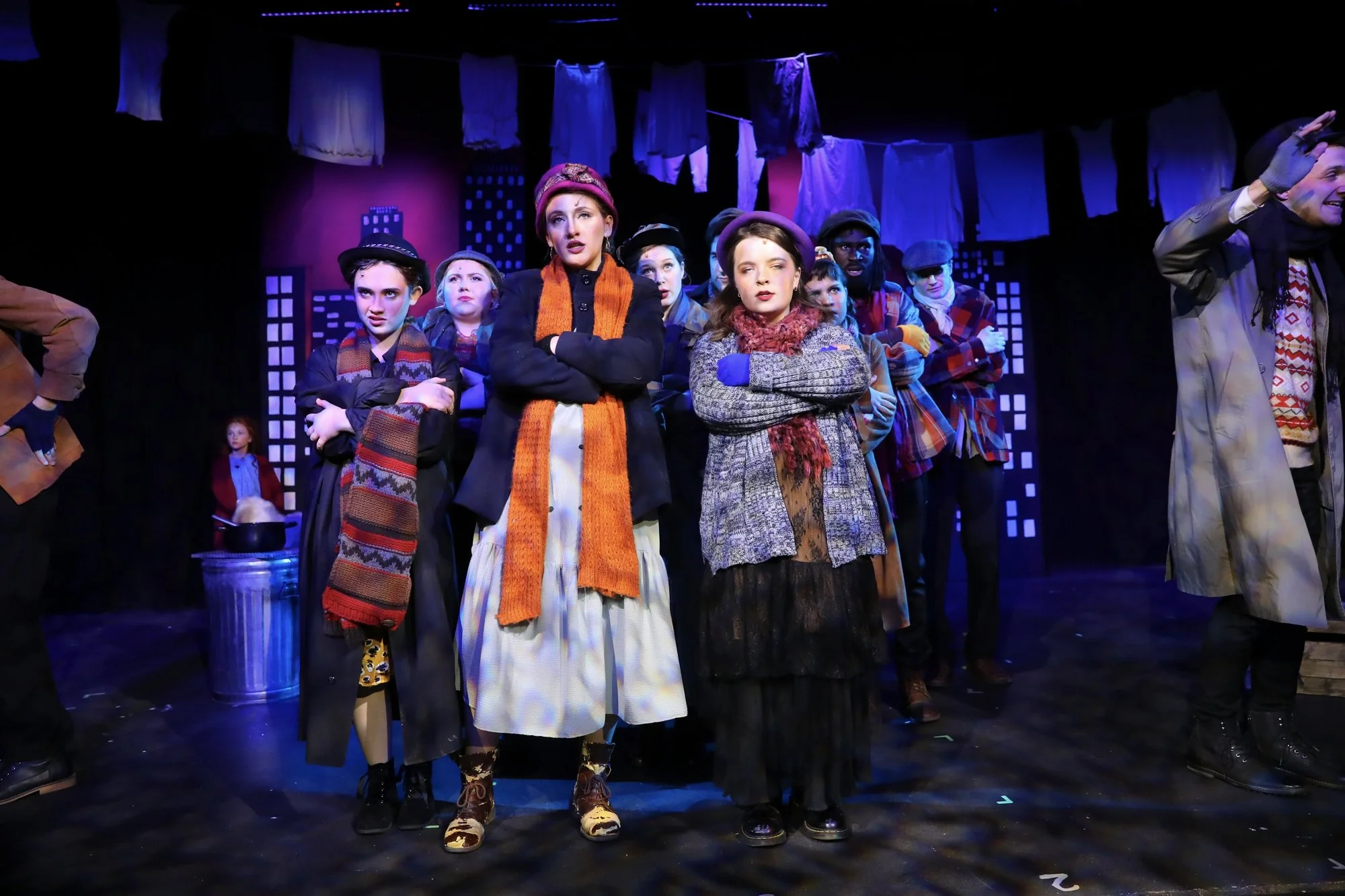 Actors on stage in a theatrical production, dressed in vintage clothing, with laundry hanging in the background, expression of concern or confusion.