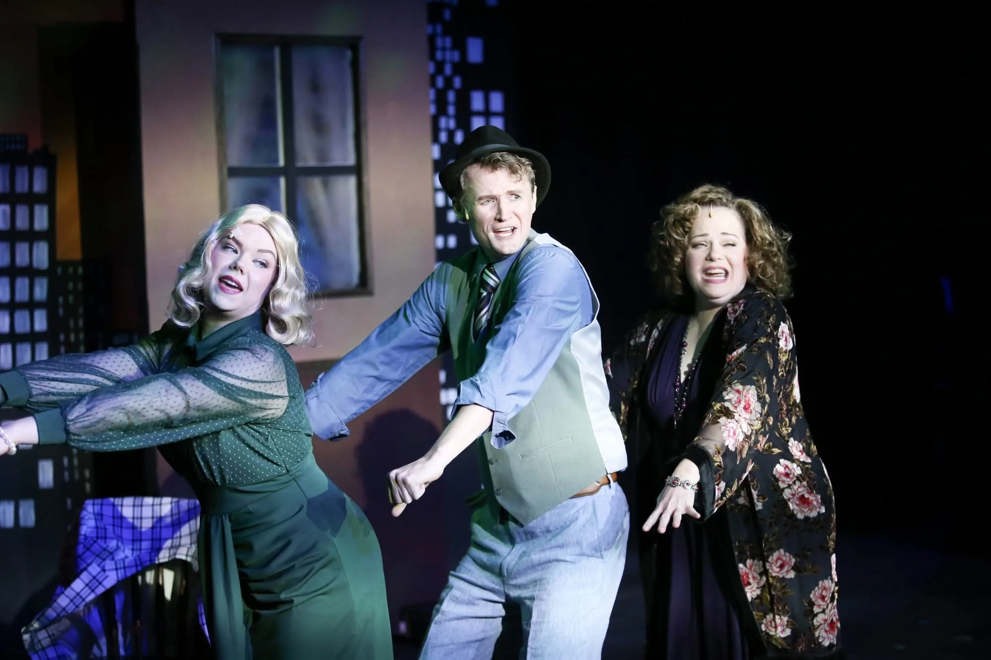 Three performers on stage in a theatrical scene, with cityscape backdrop. The performers are holding hands, expressing dramatized emotions.