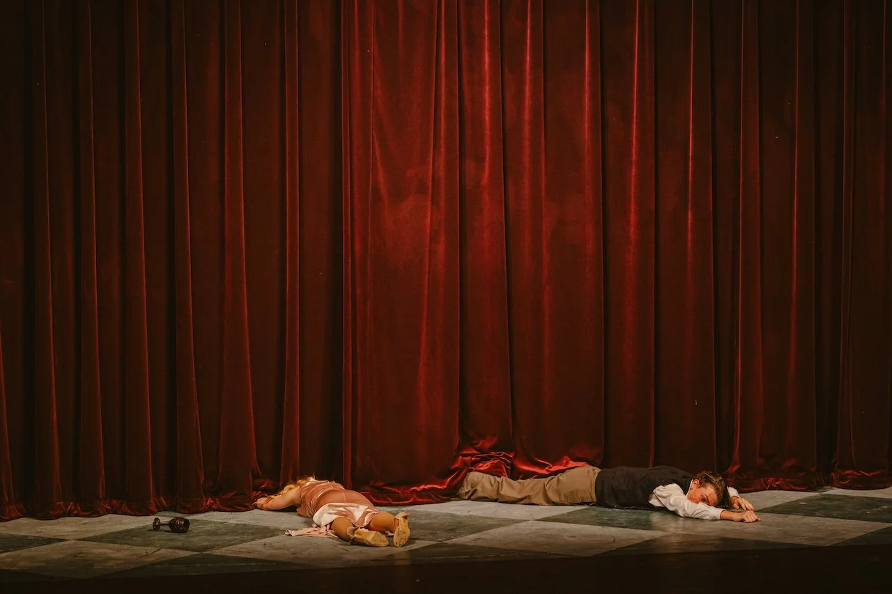Two children in theatrical costumes lying on the stage floor in front of a large red curtain, with a pair of black shoes nearby.