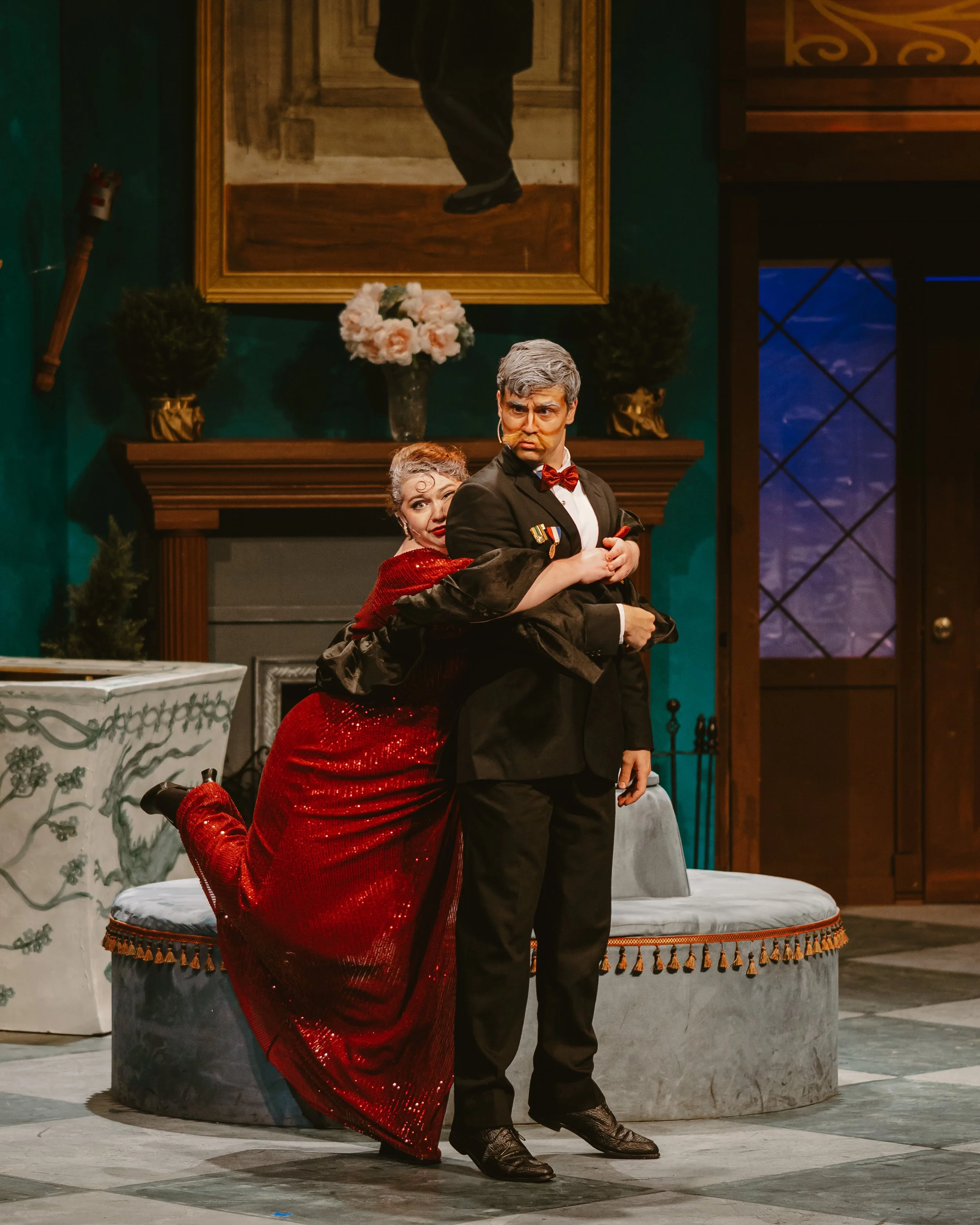 A woman and a man are performing in a theatrical play set in an ornate room. The woman is kneeling on one knee, wearing a red sequined dress and boots, with her arms wrapped around the man's waist. The man, dressed in a black tuxedo with medals and a