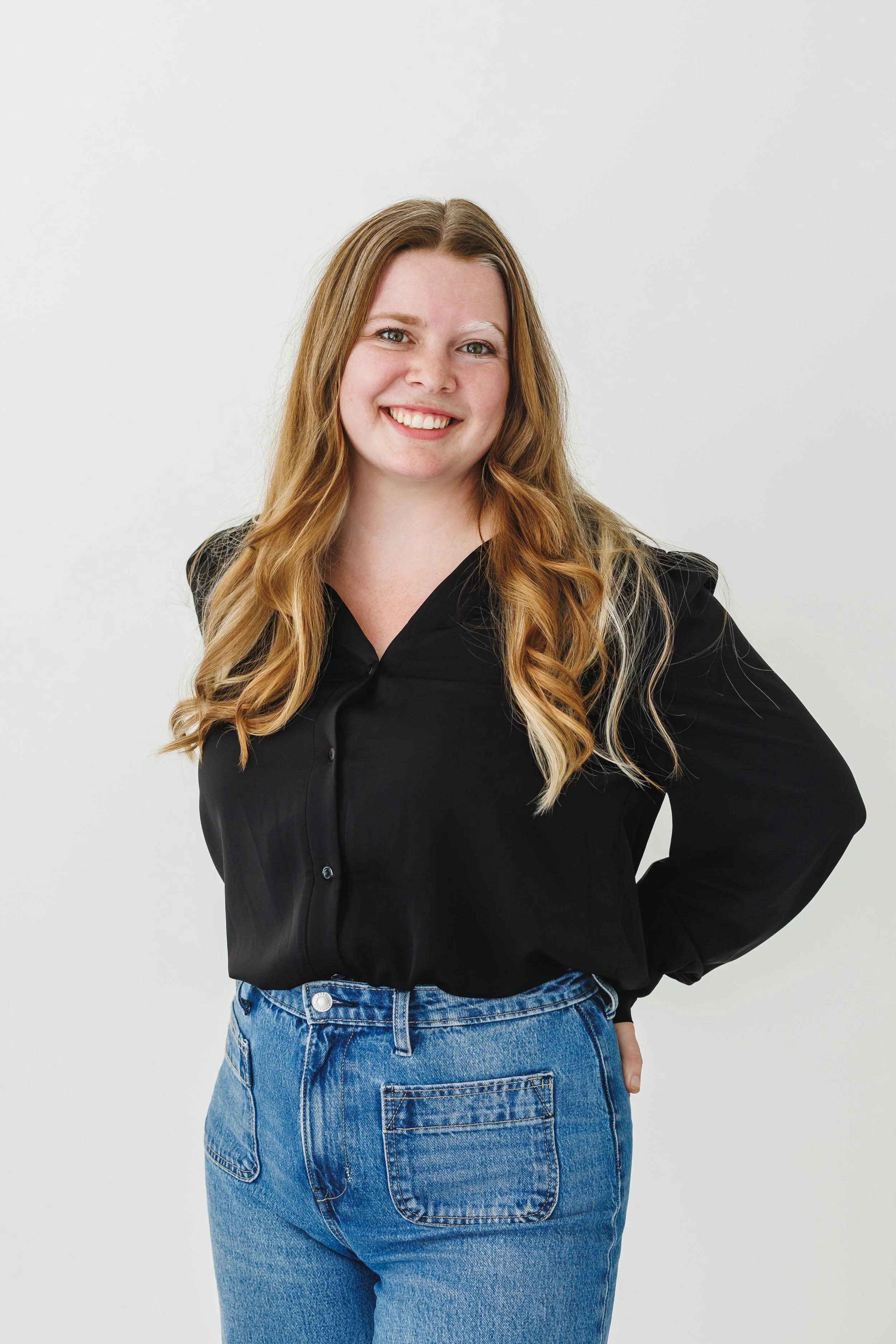A woman with long, wavy blonde hair smiling, wearing a black shirt and blue jeans, standing against a plain white background.