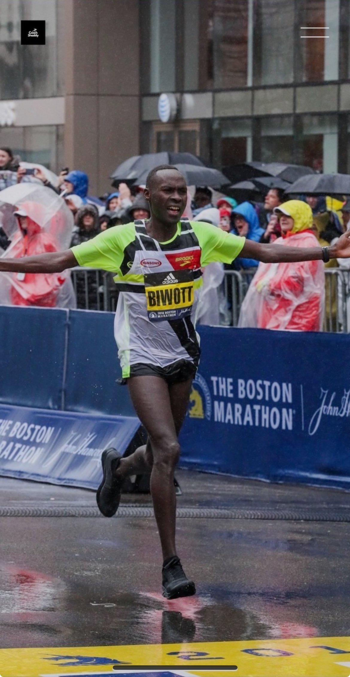 A male marathon runner wearing a yellow and black athletic shirt with a bib labeled "BIWOTT" crosses the finish line in the rain at the Boston Marathon, with spectators holding umbrellas behind metal barriers.