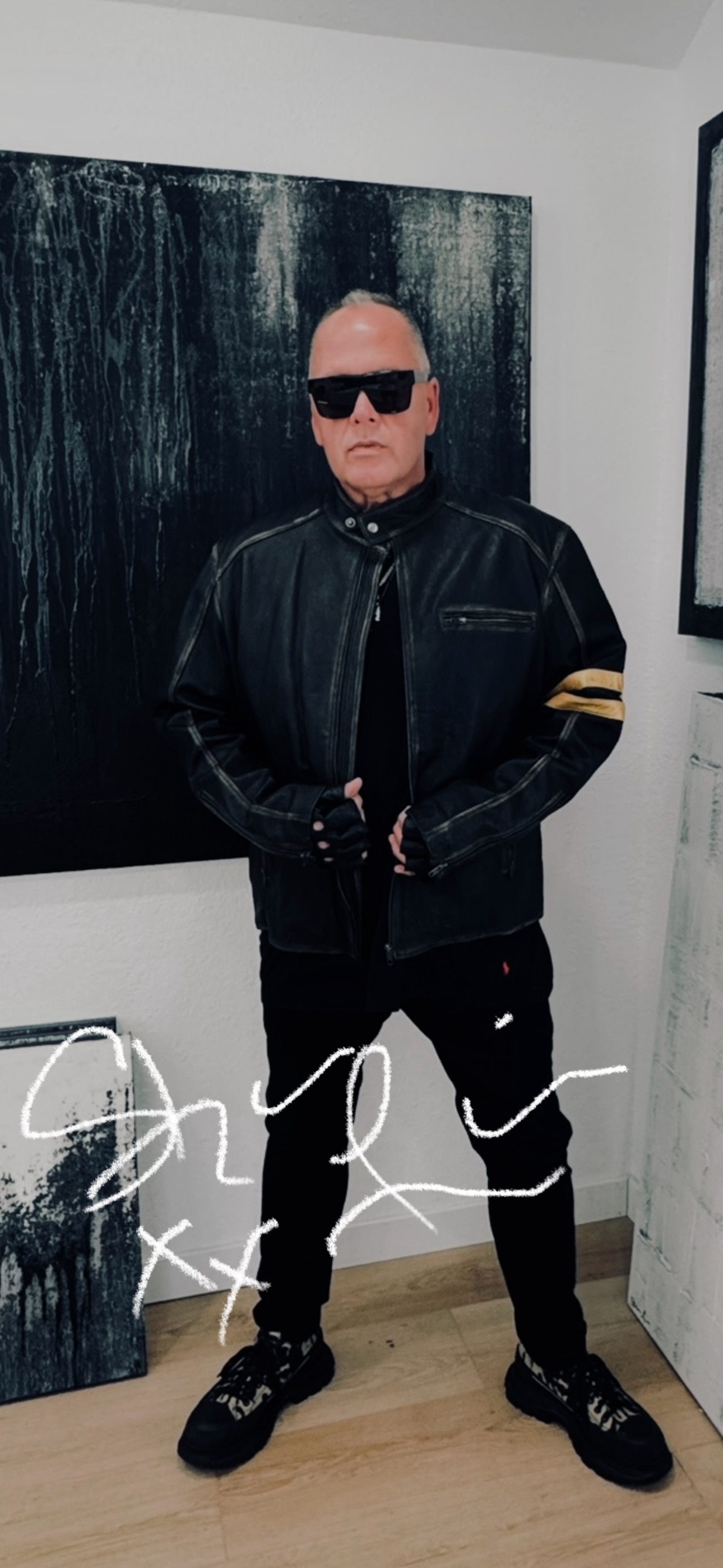 A man wearing sunglasses, a black leather jacket with a yellow stripe on the sleeve, black gloves, black pants, and black sneakers with a white pattern, standing in an art gallery with black and white abstract paintings.