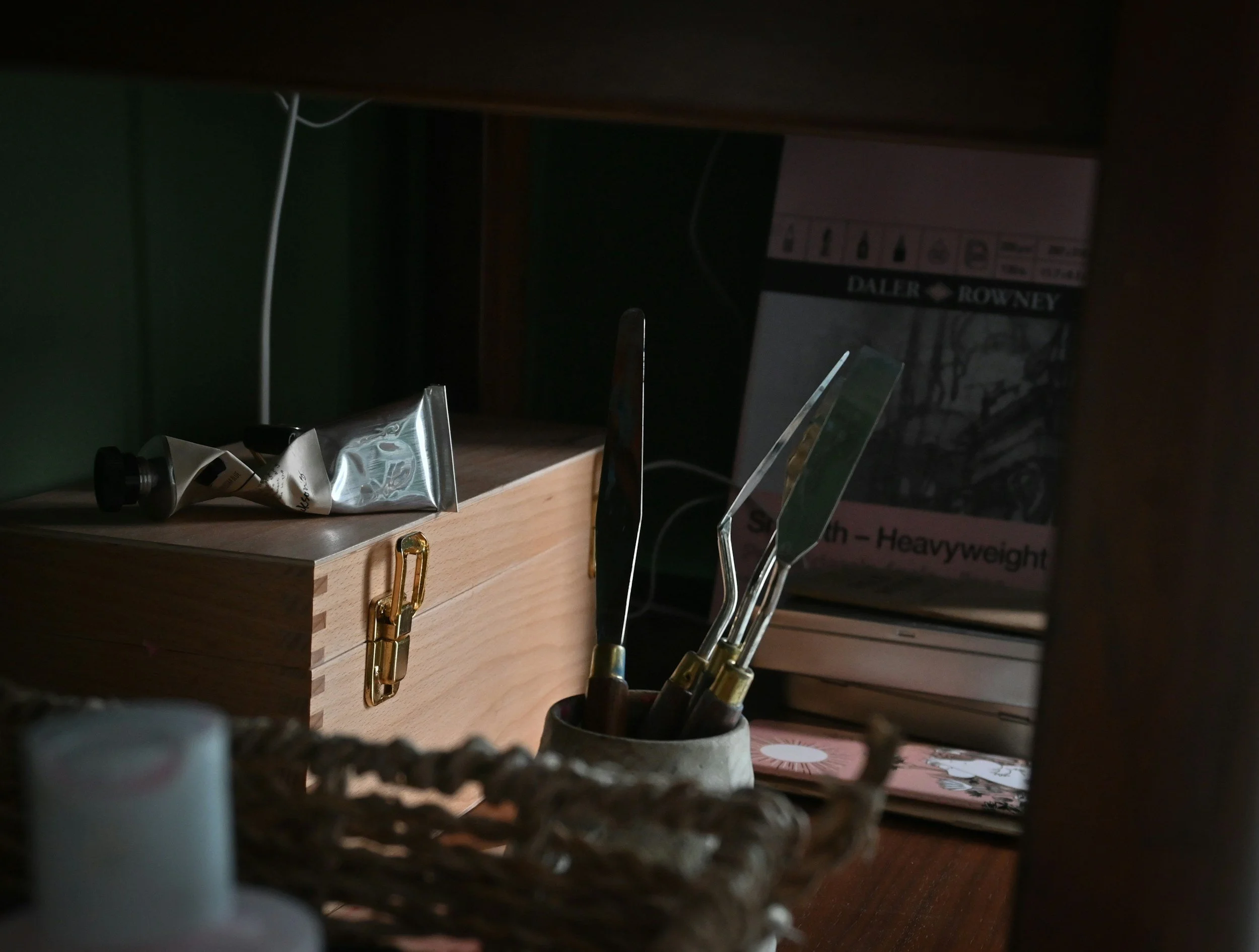 A wooden desk or shelf with art tools, including carving or sculpting knives, stored in a small container. A Lego set box is visible in the background.