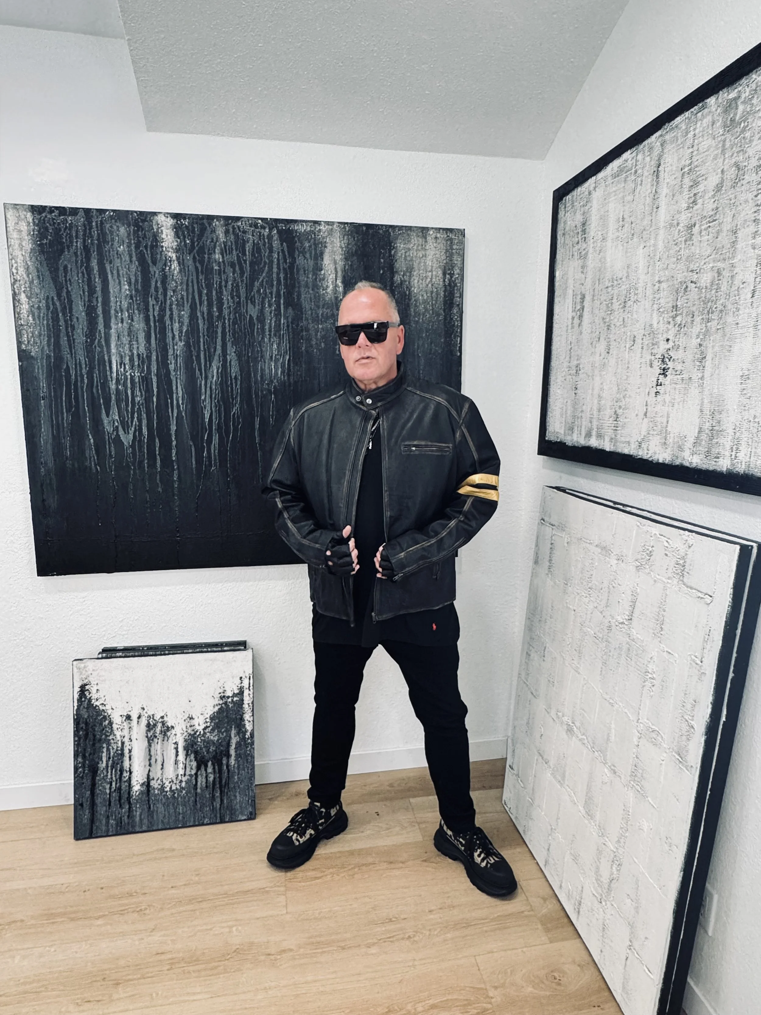 A man wearing sunglasses and a black leather jacket standing inside an art gallery, surrounded by black and white abstract paintings.