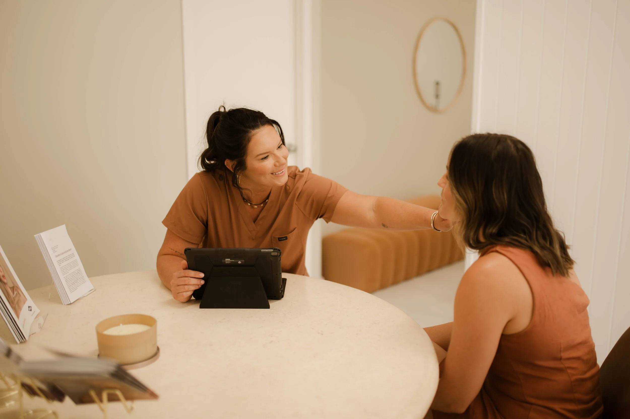 A woman in medical scrubs smiling and reaching out to comfort a seated woman in a waiting area.