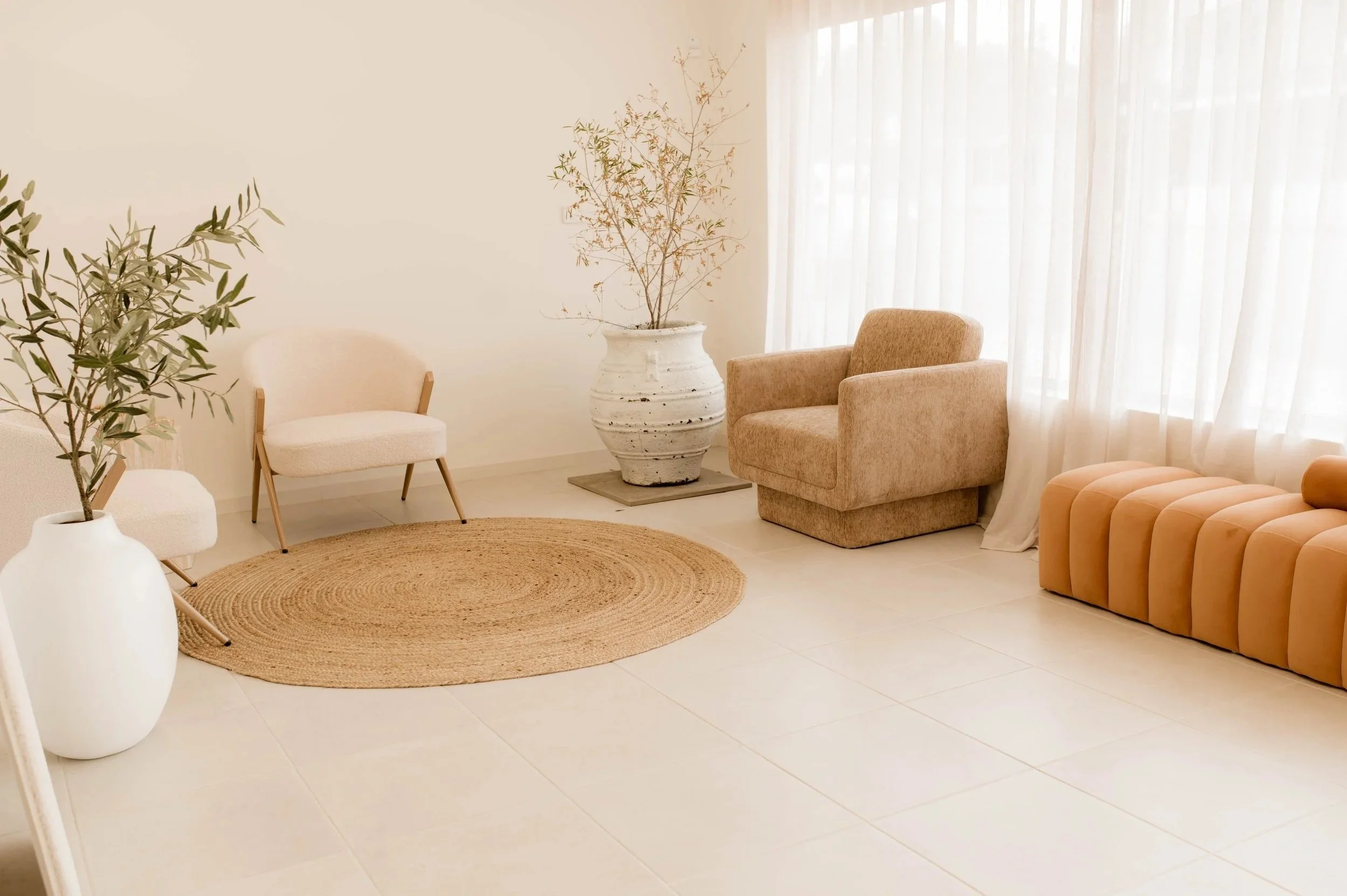 Minimalist waiting room with beige armchair, tan ottoman, two white chairs, large potted plants, round jute rug, and sheer white curtains.