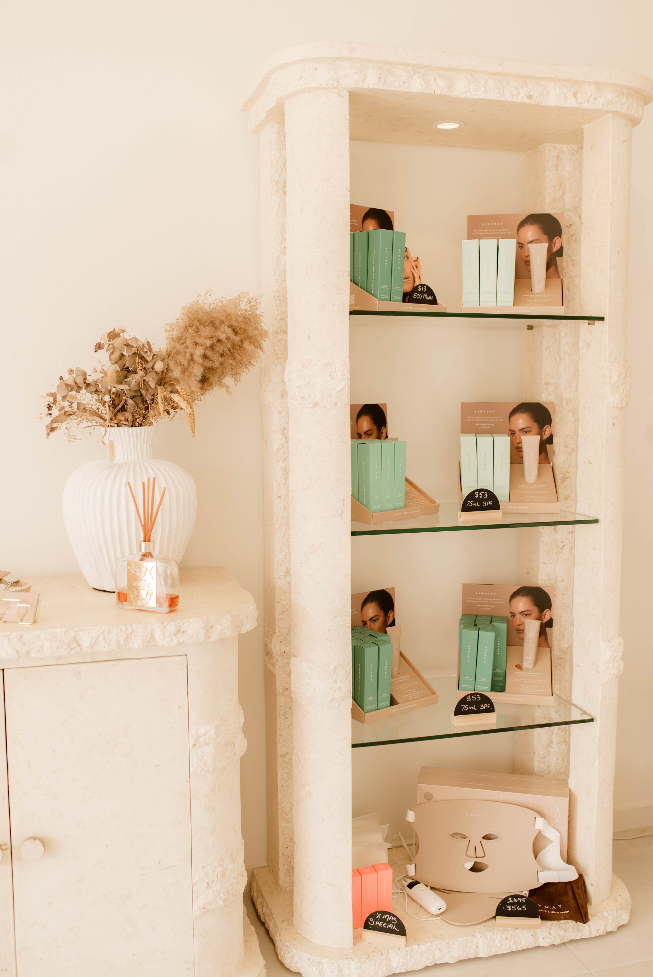Display shelf with skincare products, a vase with dried flowers, and a diffuser in a neutral-colored, spa-like setting.