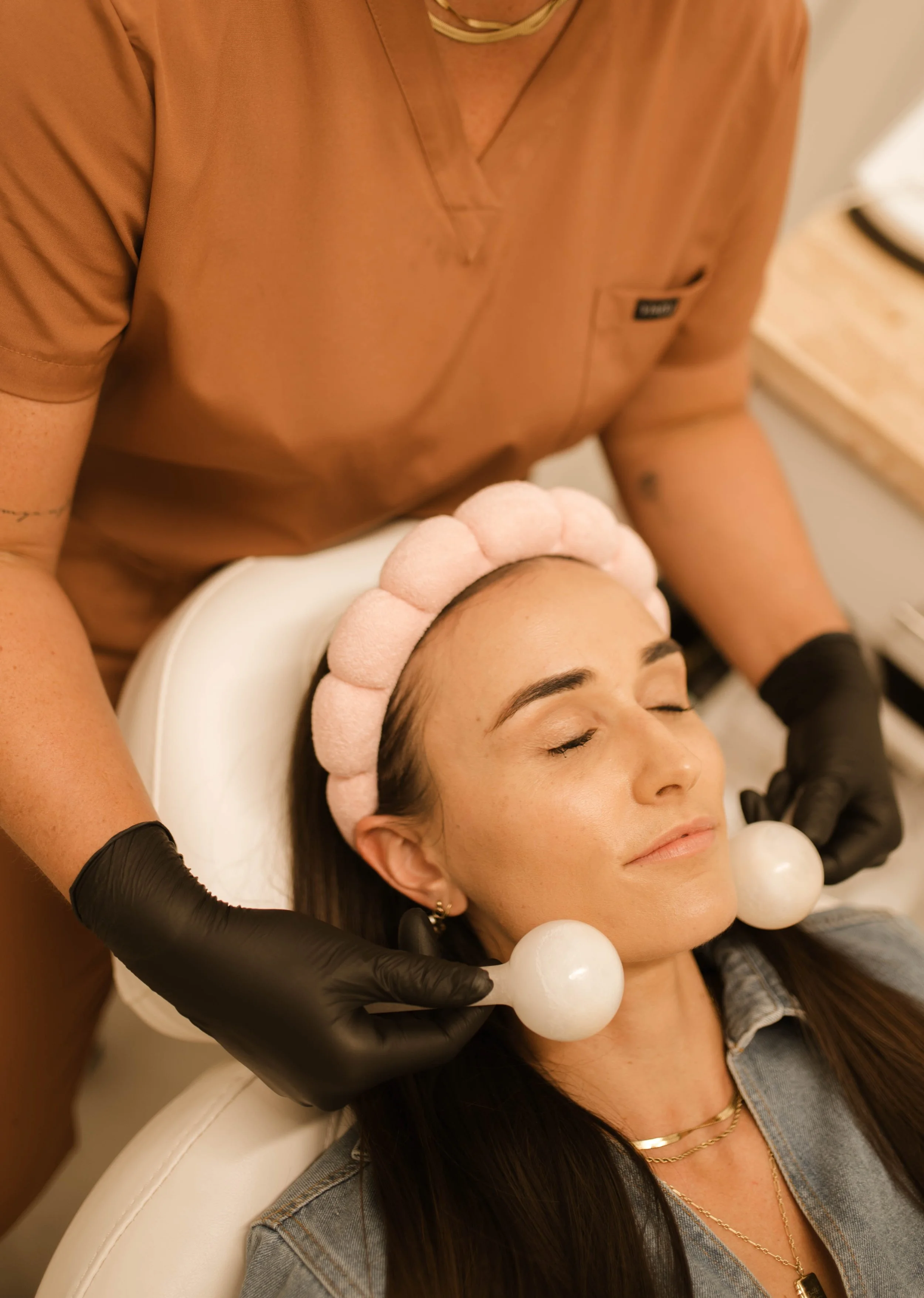 A woman receiving a facial treatment at a spa, with her eyes closed and a pink towel headband, while a technician in black gloves holds two massage tools next to her face.