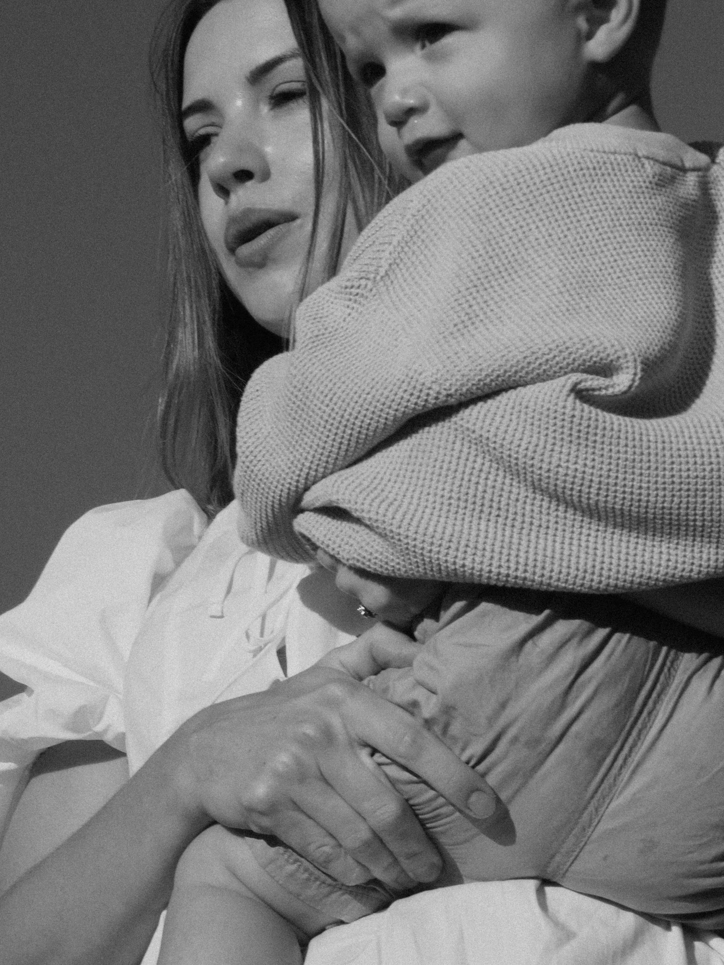 Black and white photo of a woman holding a baby, both looking into the distance. The baby is wrapped in a textured blanket.