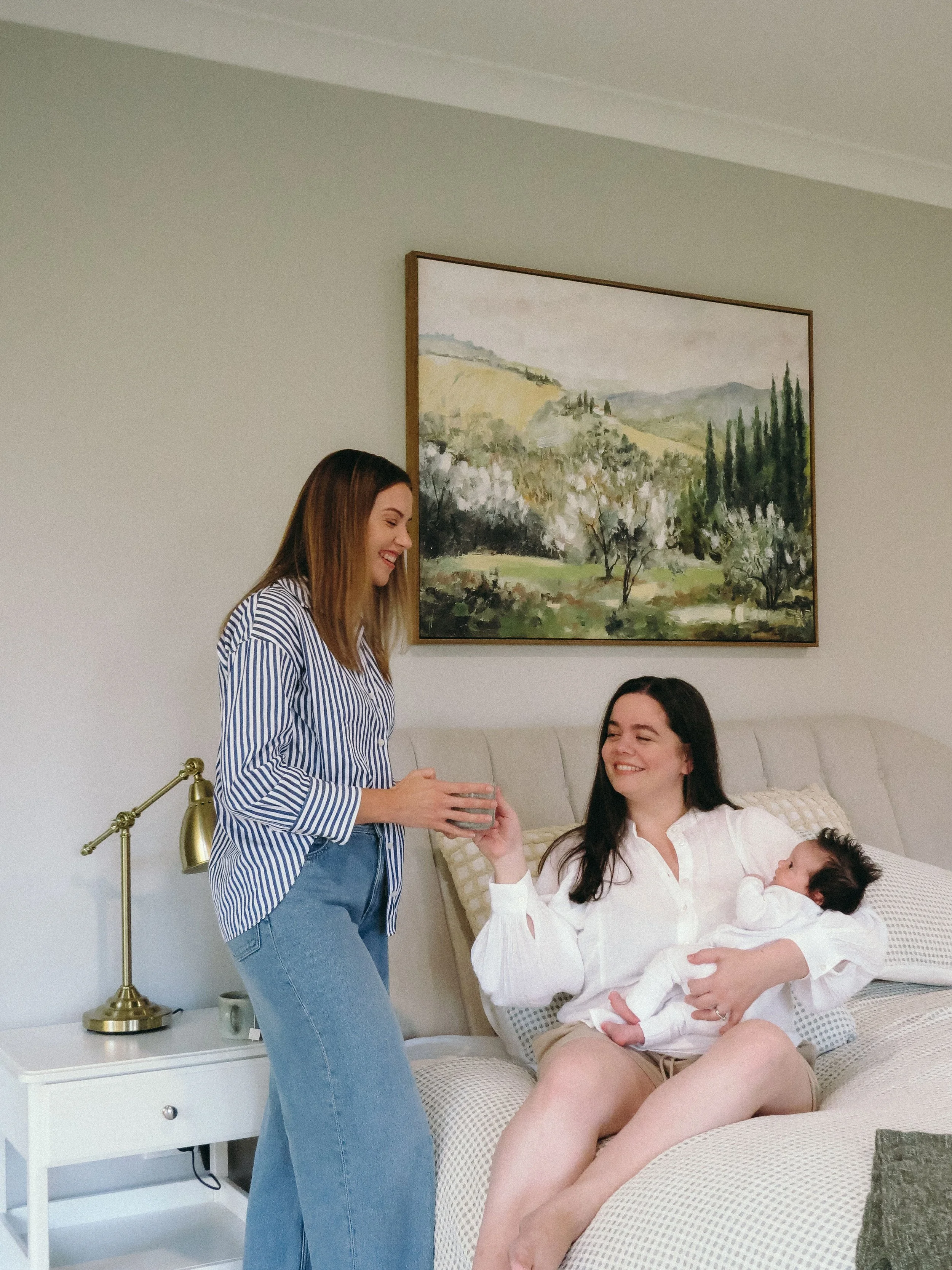 Woman sitting on bed holding baby, receiving a drink from another woman standing; cozy bedroom setting with landscape painting on wall.