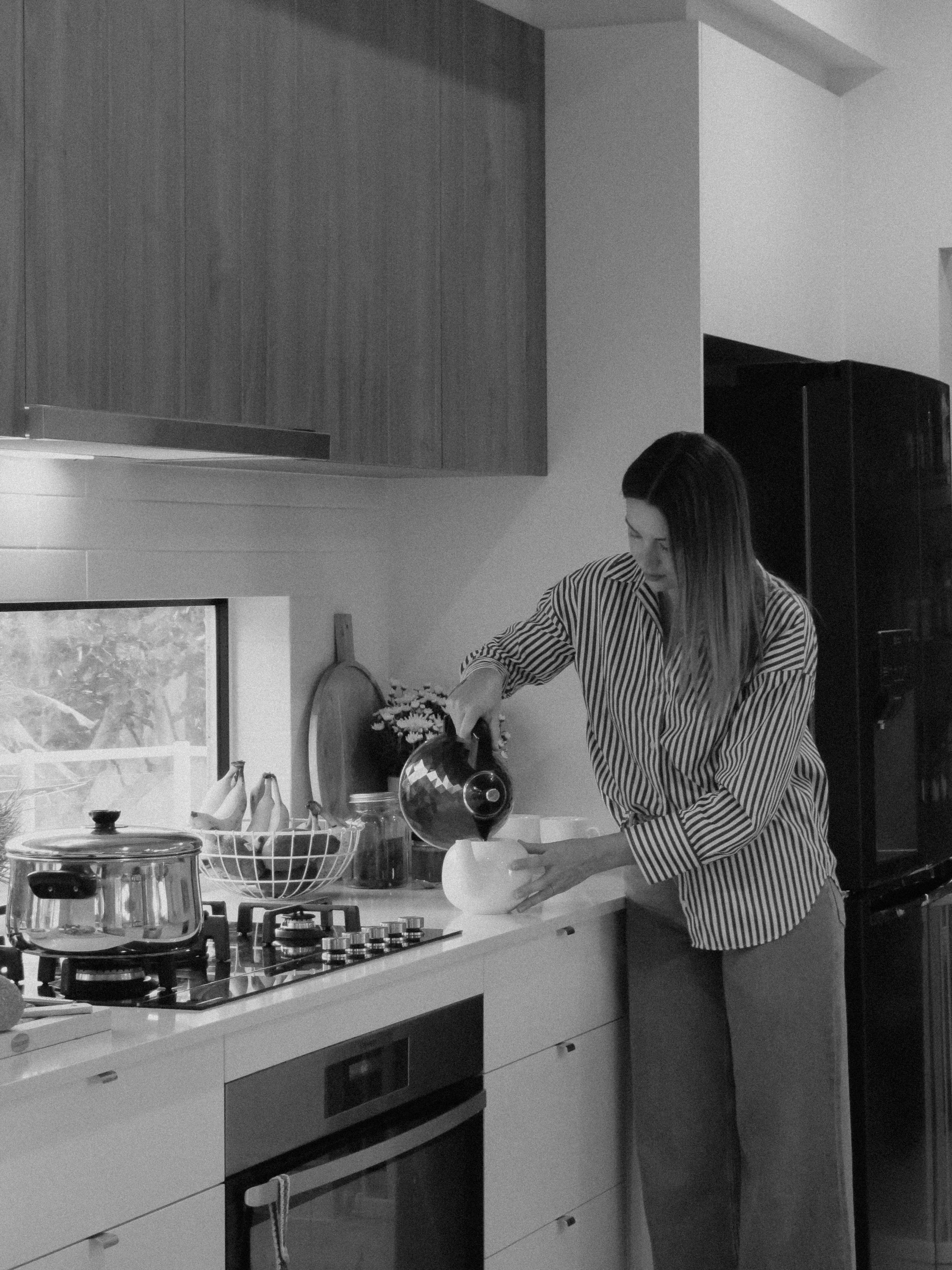 Woman in a kitchen pouring coffee into a mug, with a stovetop, pots, and kitchen appliances visible.