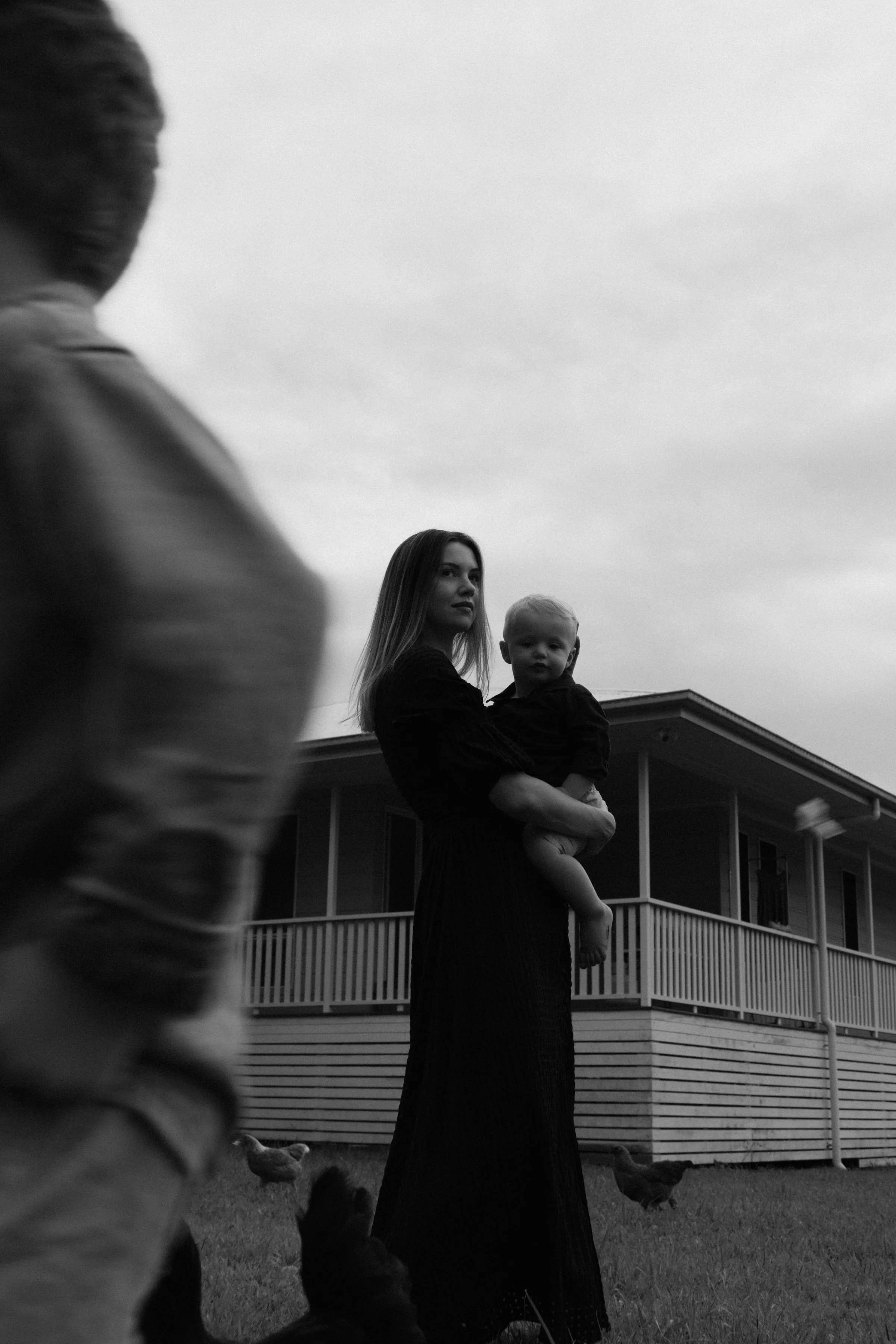Black and white photo of a woman holding a baby in front of a house with a porch, with a blurred child in the foreground and chickens on the lawn.