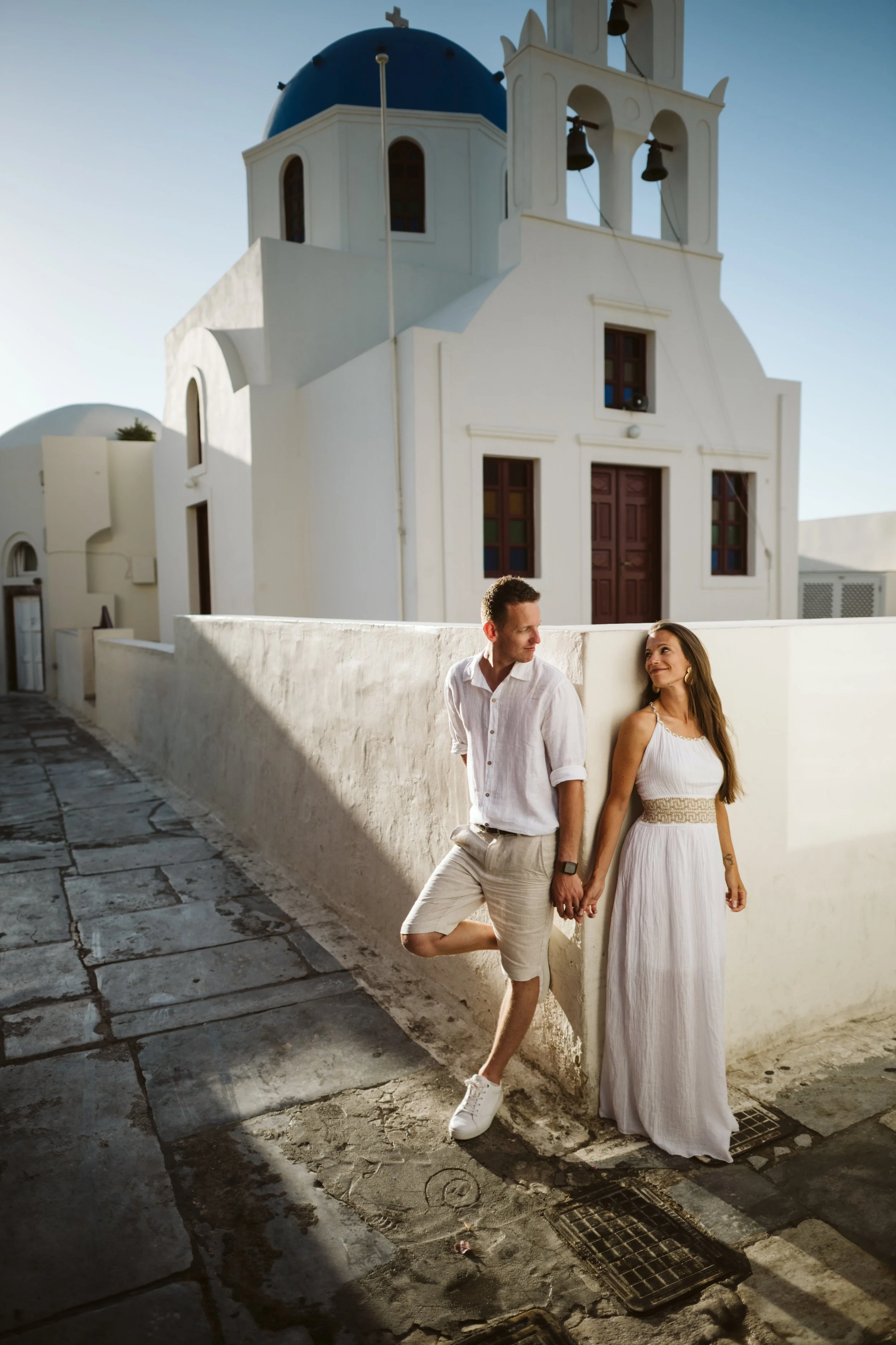 A couple holding hands and smiling on a sidewalk with a white building and blue-domed roof in the background.
