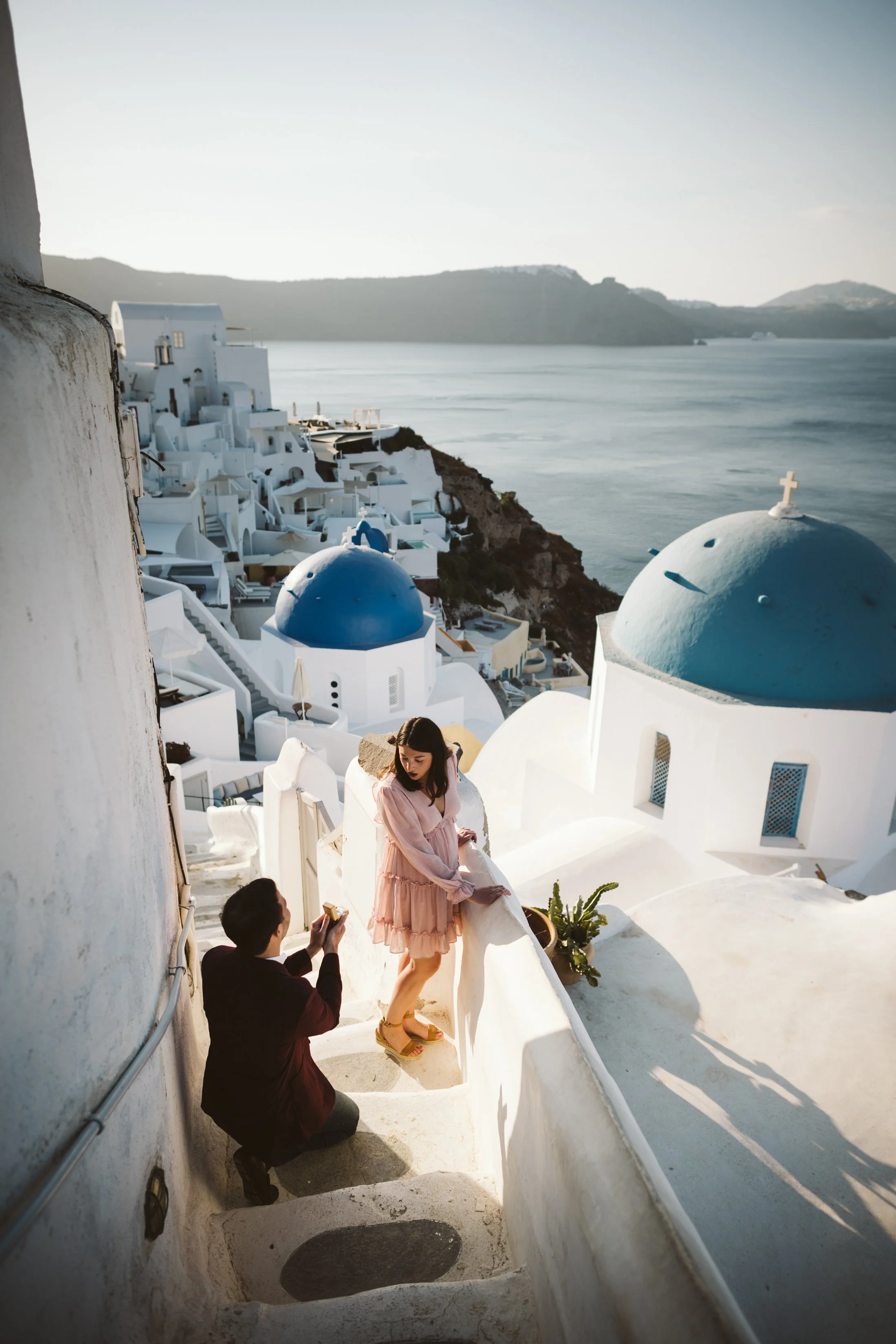 A woman in a pink dress and sandals is standing on a white curved staircase in Santorini, Greece, with a man in a dark suit kneeling and taking a photo of her. The background shows white buildings with blue domes and the Aegean Sea.