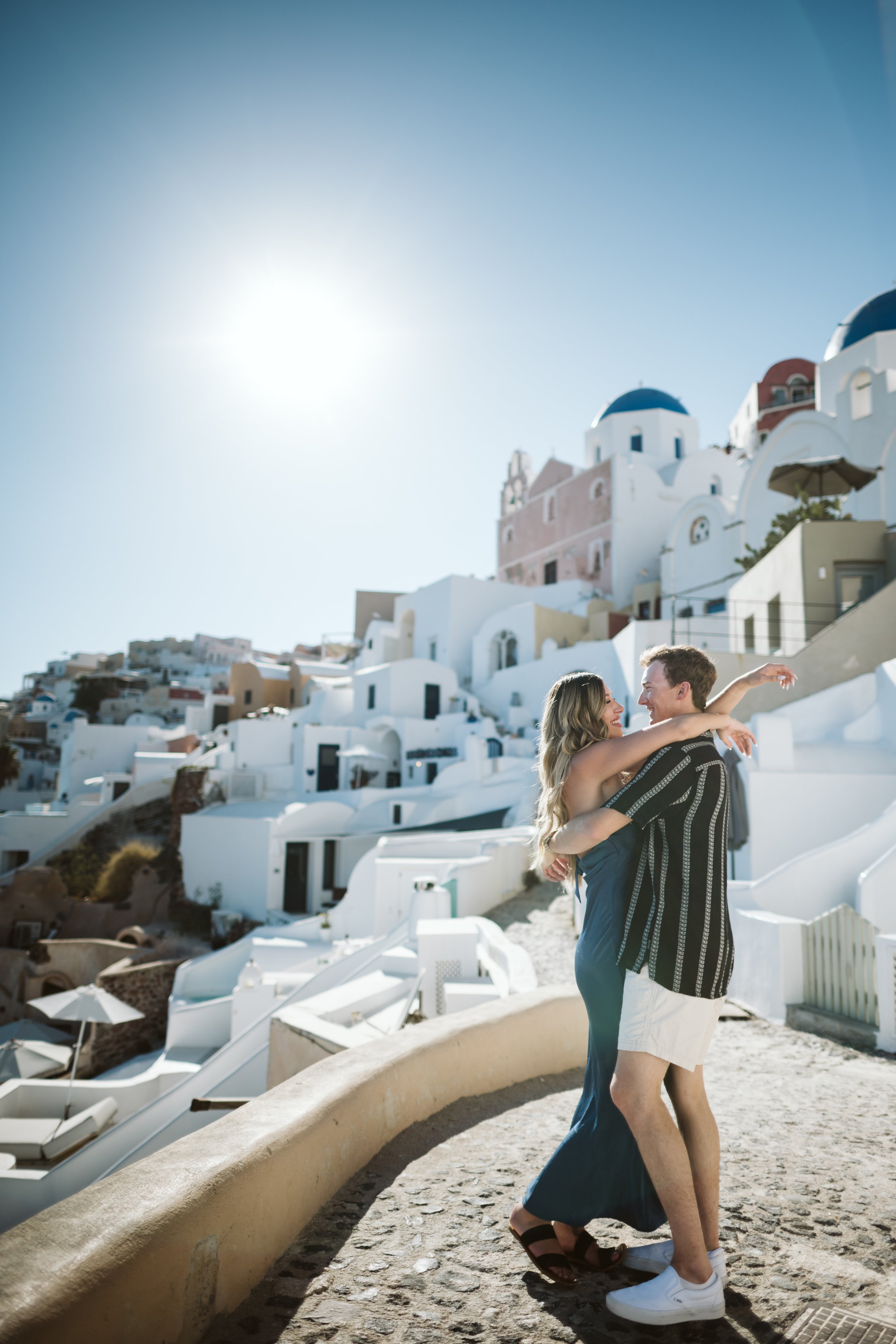 A young couple dancing outdoors near white buildings with domed roofs in Santorini, Greece, on a sunny day.