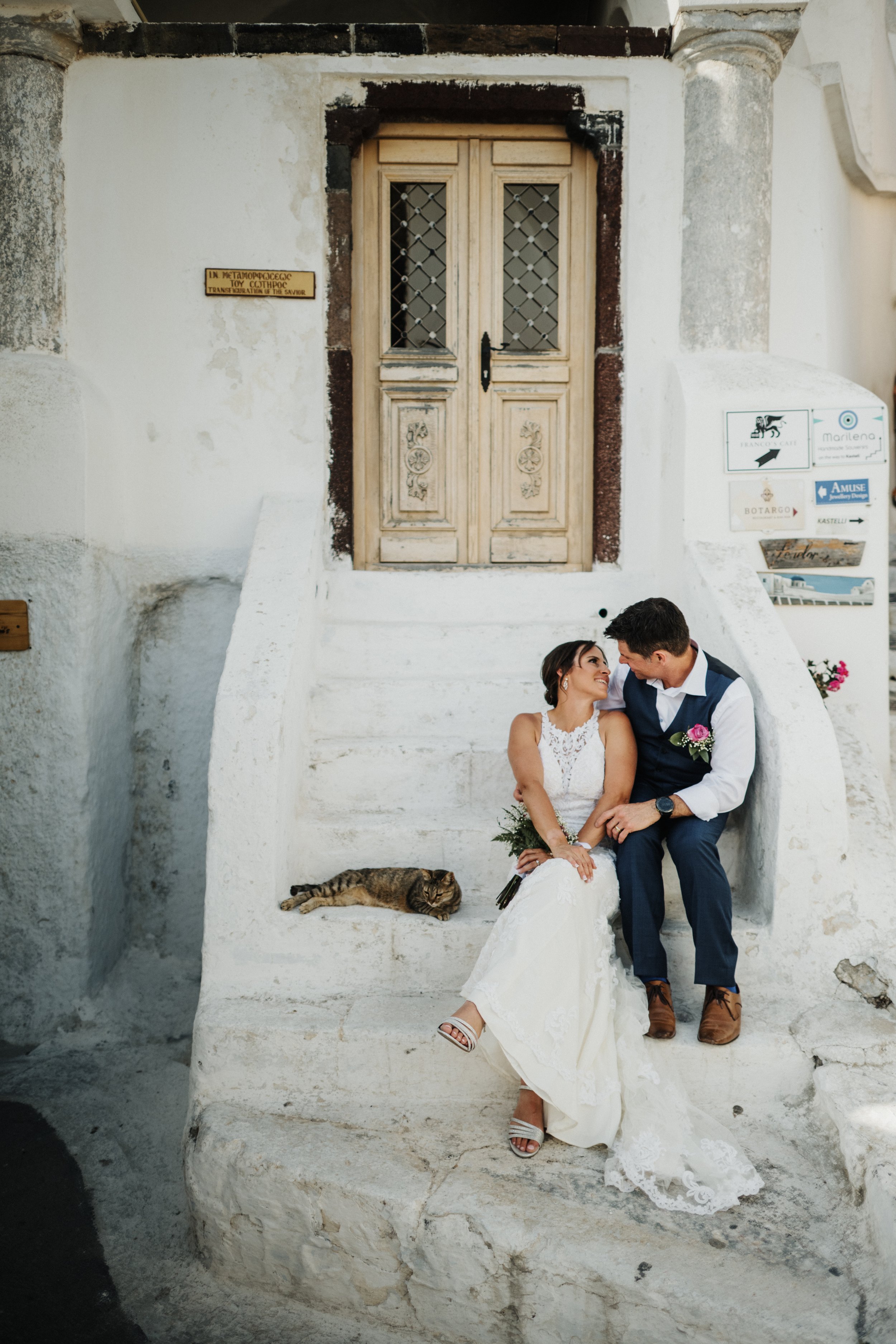 A bride and groom sitting on stone steps in front of a rustic wooden door with decorative glass panels, smiling at each other, with a cat lying on the step beside them.