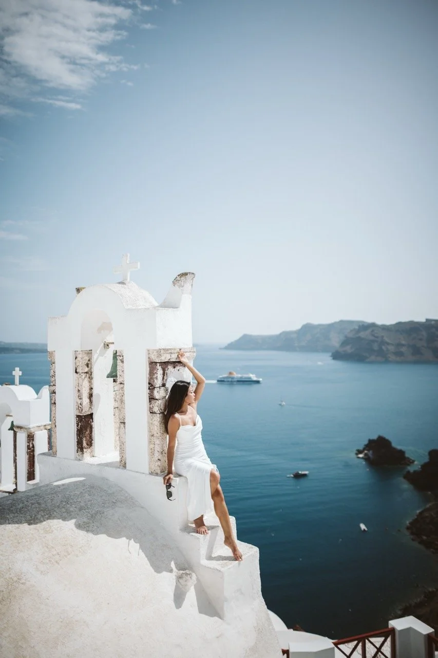A woman in a white dress sitting on a ledge of a white building with a cross on top, overlooking a blue sea with boats and cliffs in the background.