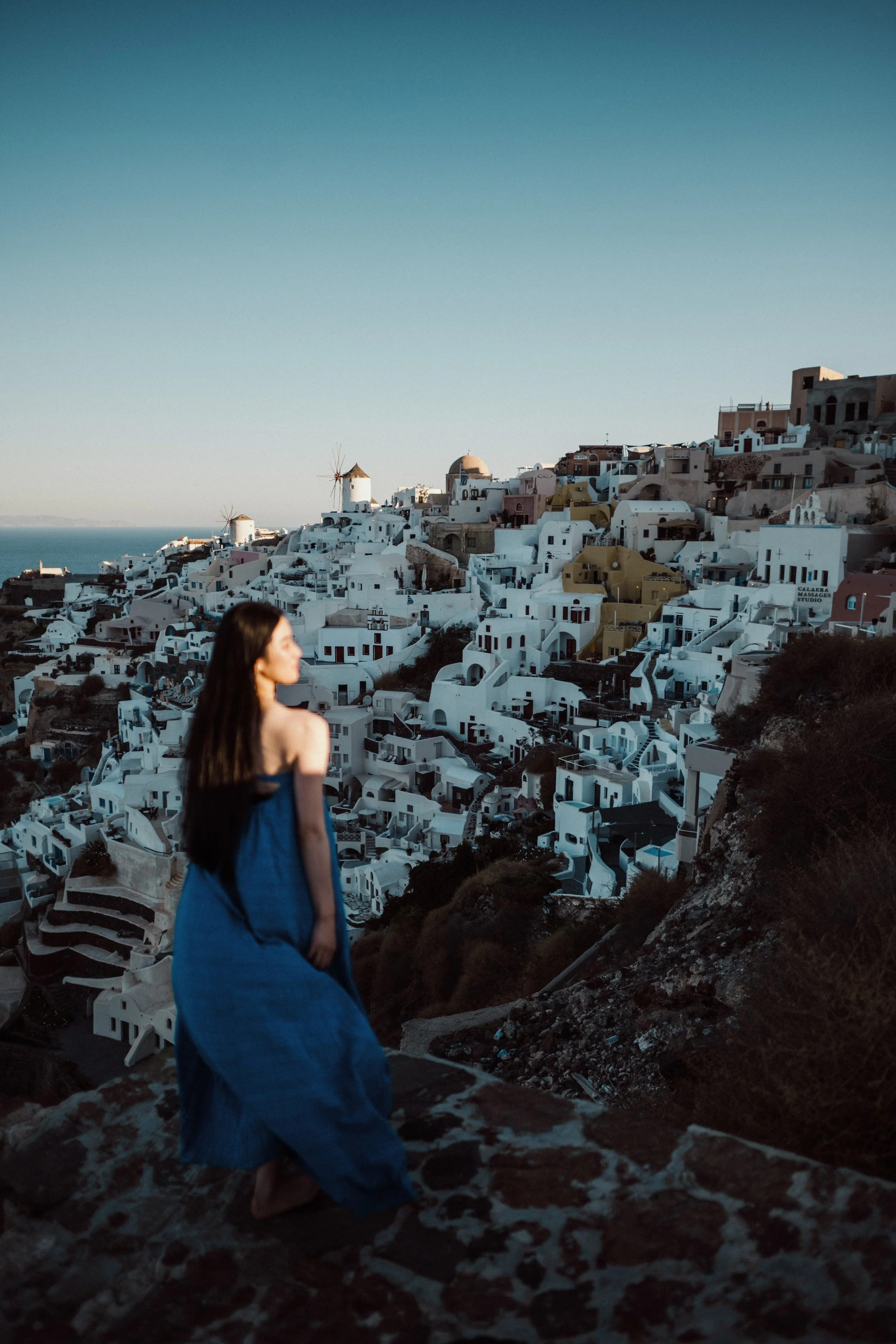 A woman with long dark hair in a blue dress stands on rocky terrain with a white hillside town featuring windmills and traditional architecture against a clear blue sky.