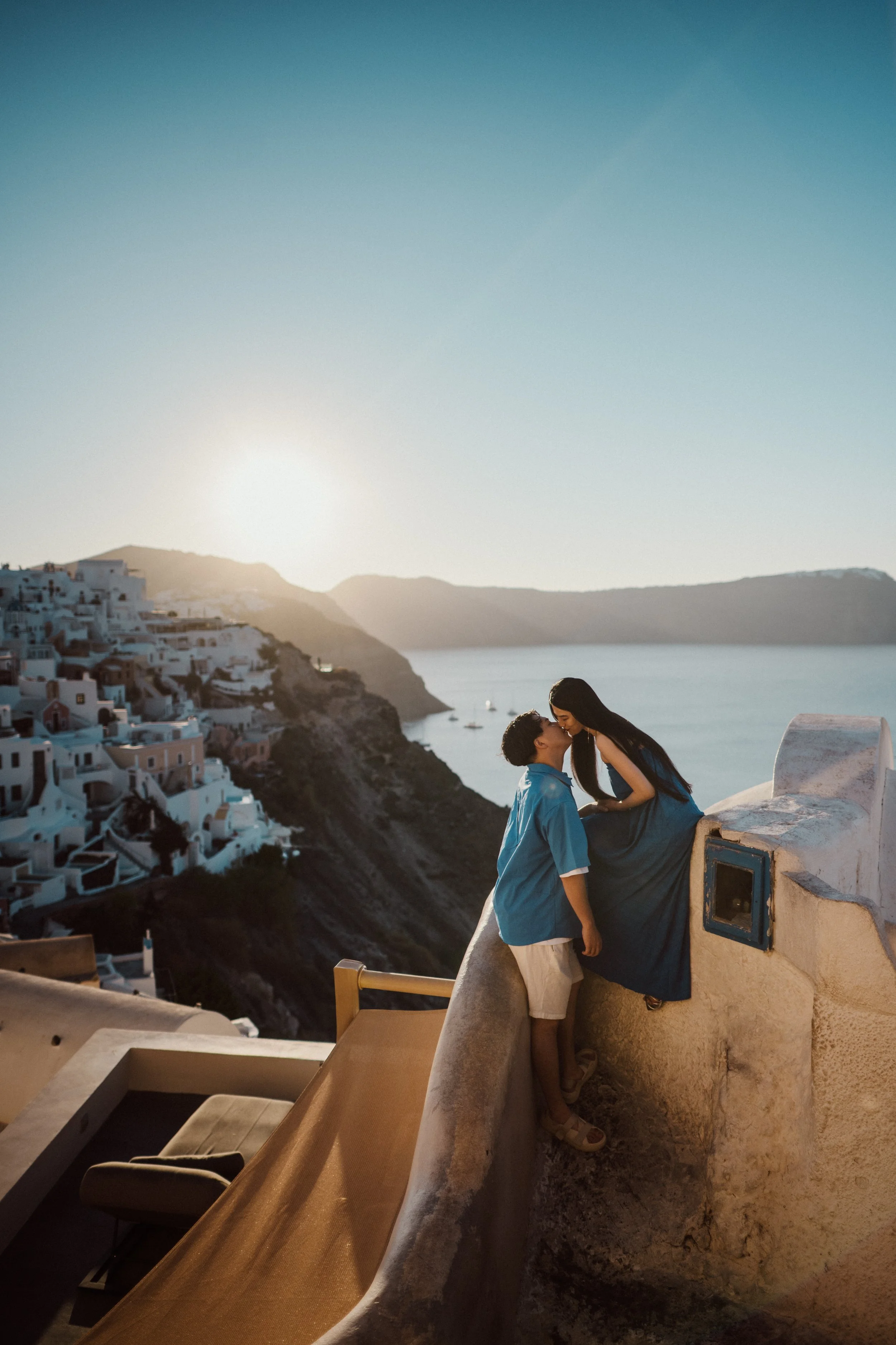 A couple standing on a rooftop, about to kiss, overlooking a coastal town with white buildings and a caldera, with the sea and distant hills in the background during sunset.