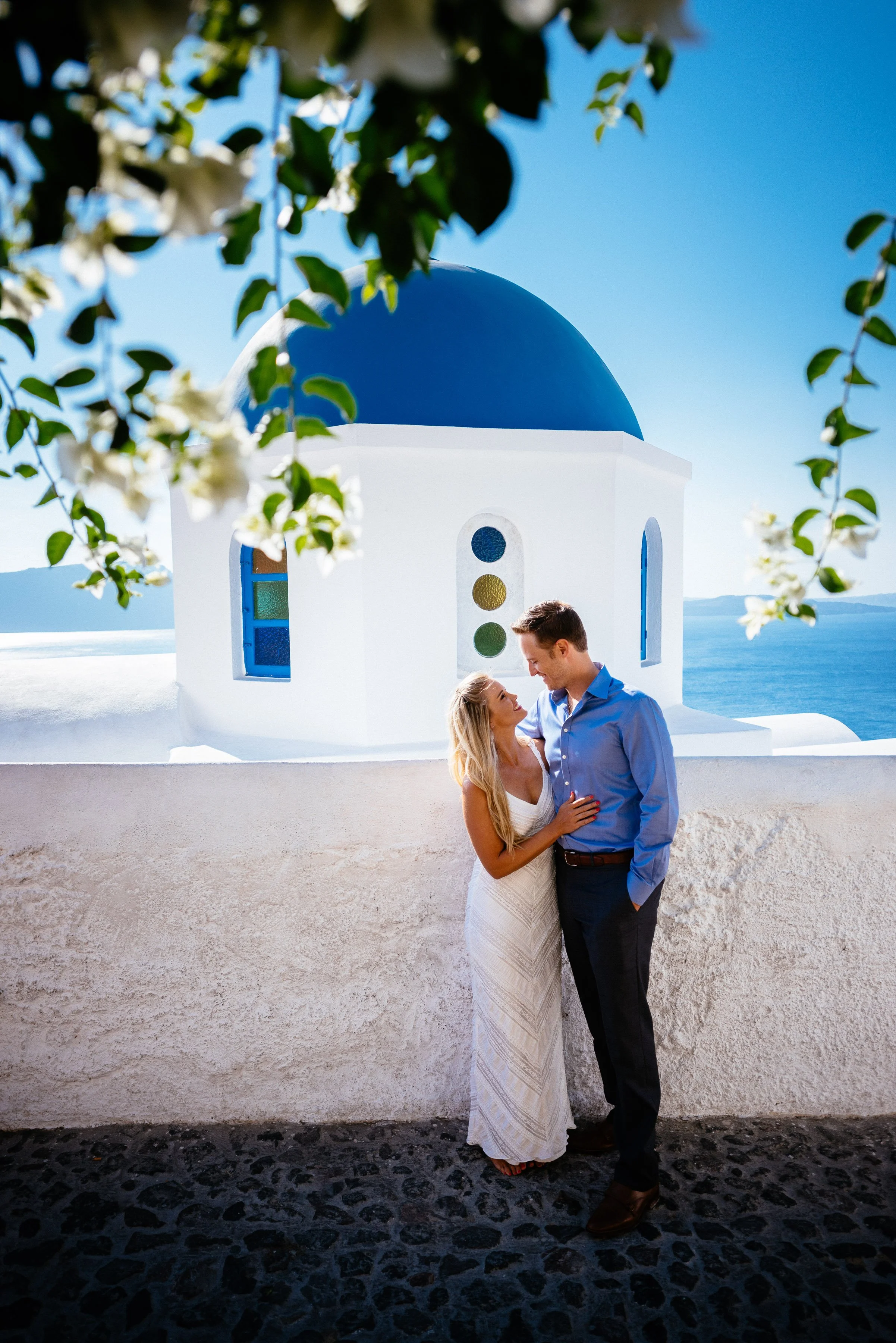 A couple standing close together near a white wall with a blue-domed church in the background, overlooking the sea on a sunny day in Greece.