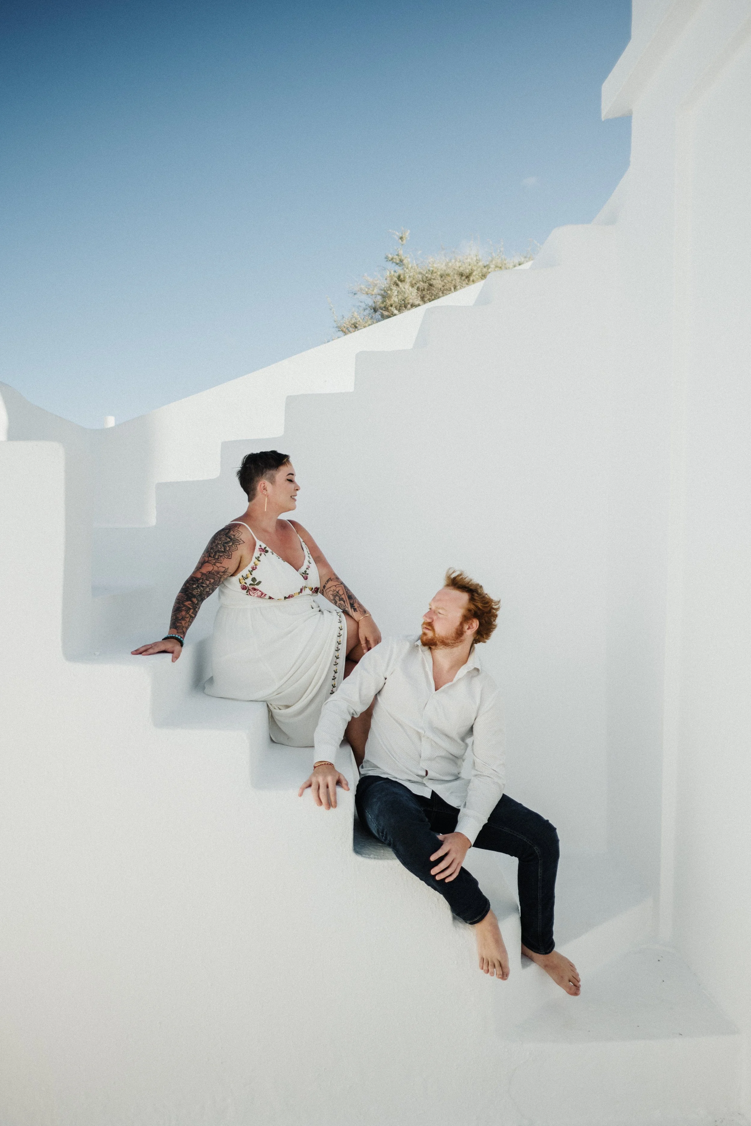 A woman with short hair and tattoos wearing a white dress with floral embroidery, and a man with a beard and short hair wearing a white shirt, sitting together on a white architectural structure with stairs and a blue sky in the background.
