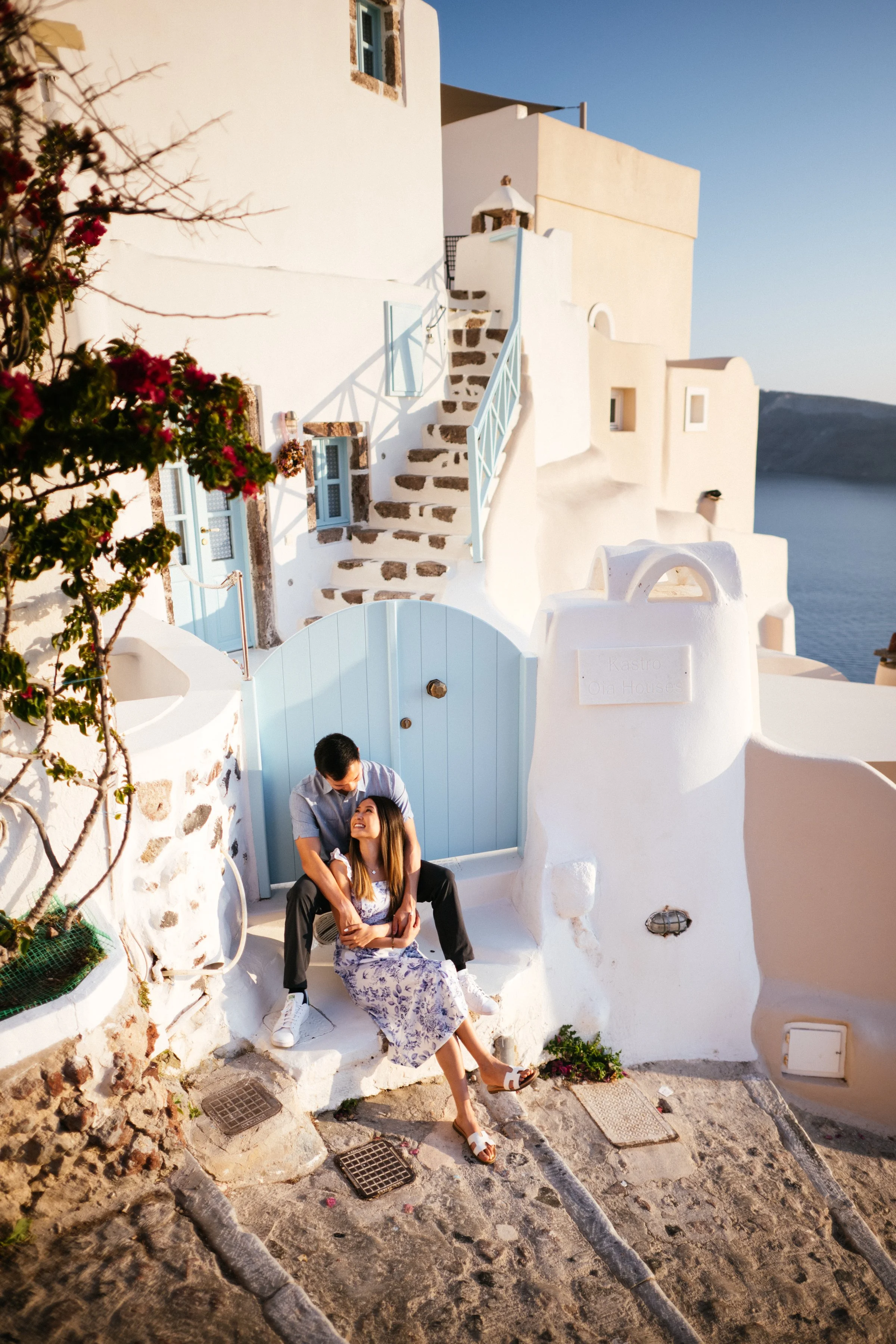 A couple sitting on a white stone staircase outside a white building with a blue door, overlooking the sea, with the man standing behind and embracing the woman, both smiling and enjoying the sunset.