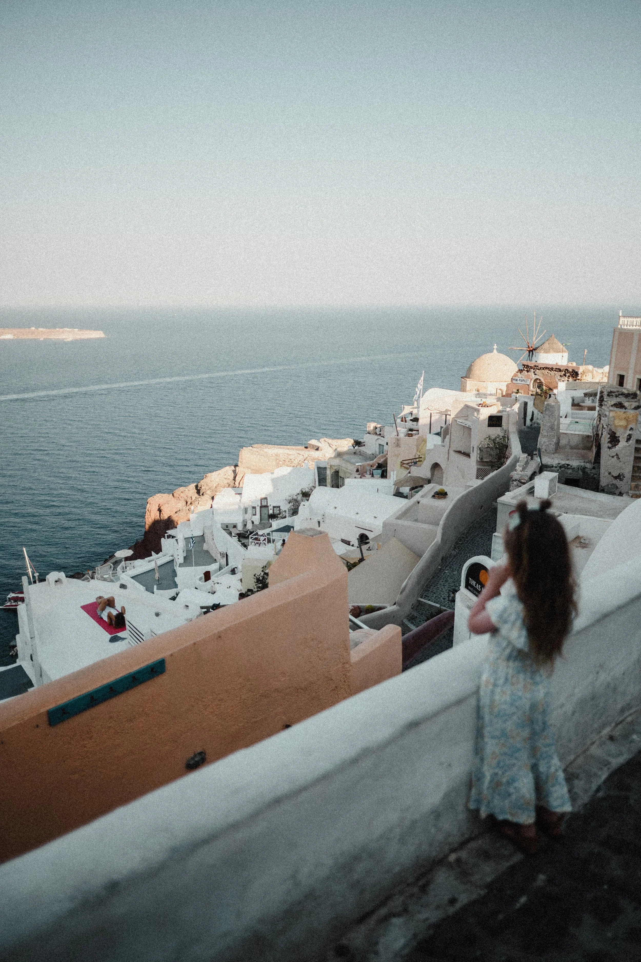 A girl in a floral dress standing on a balcony overlooking white buildings and the ocean in Santorini, Greece, during daytime.