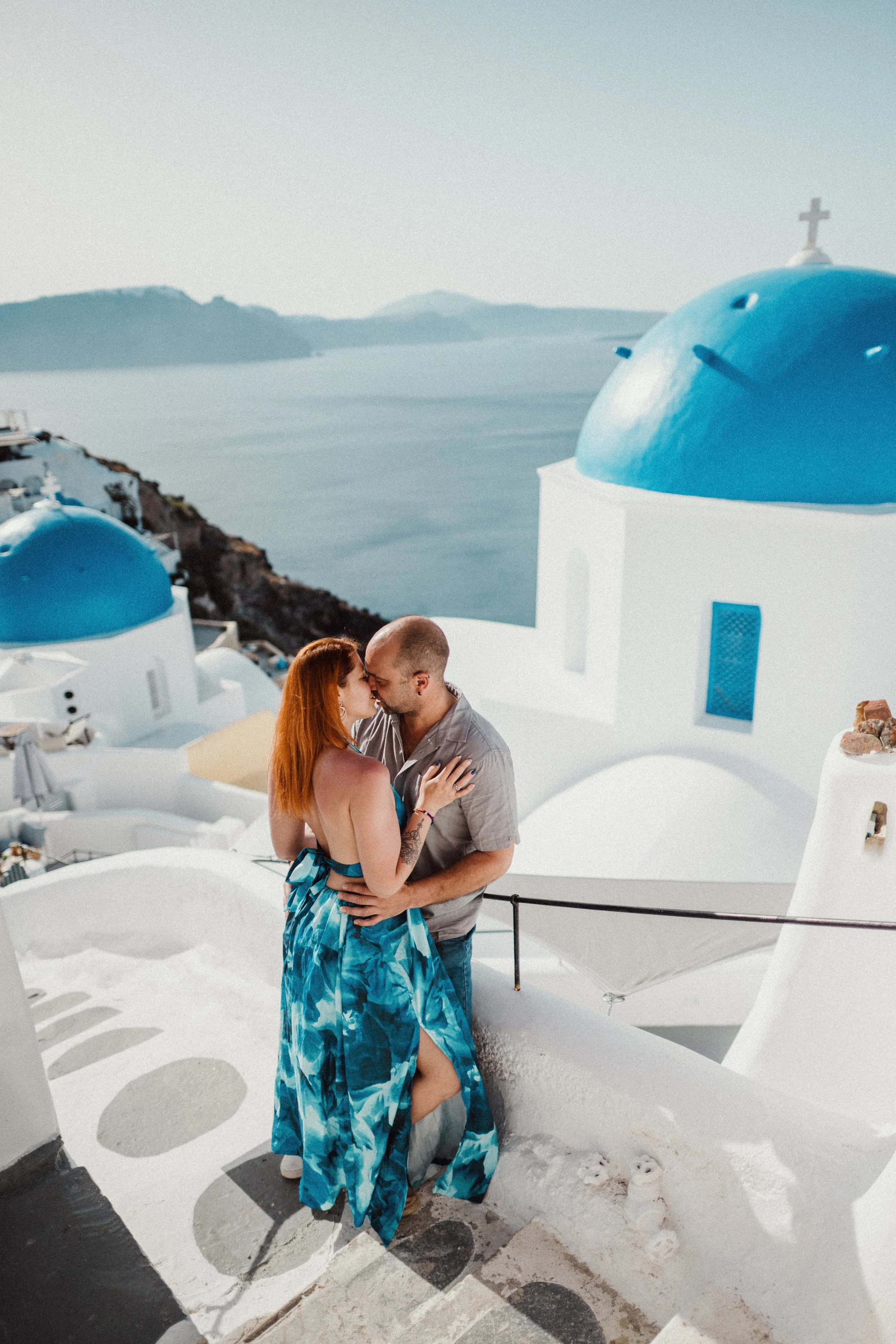A couple is standing close together on a white-painted pavement in Santorini, Greece. The woman has long red hair, and the man is bald. They are touching foreheads and looking at each other affectionately. In the background, white buildings with blue