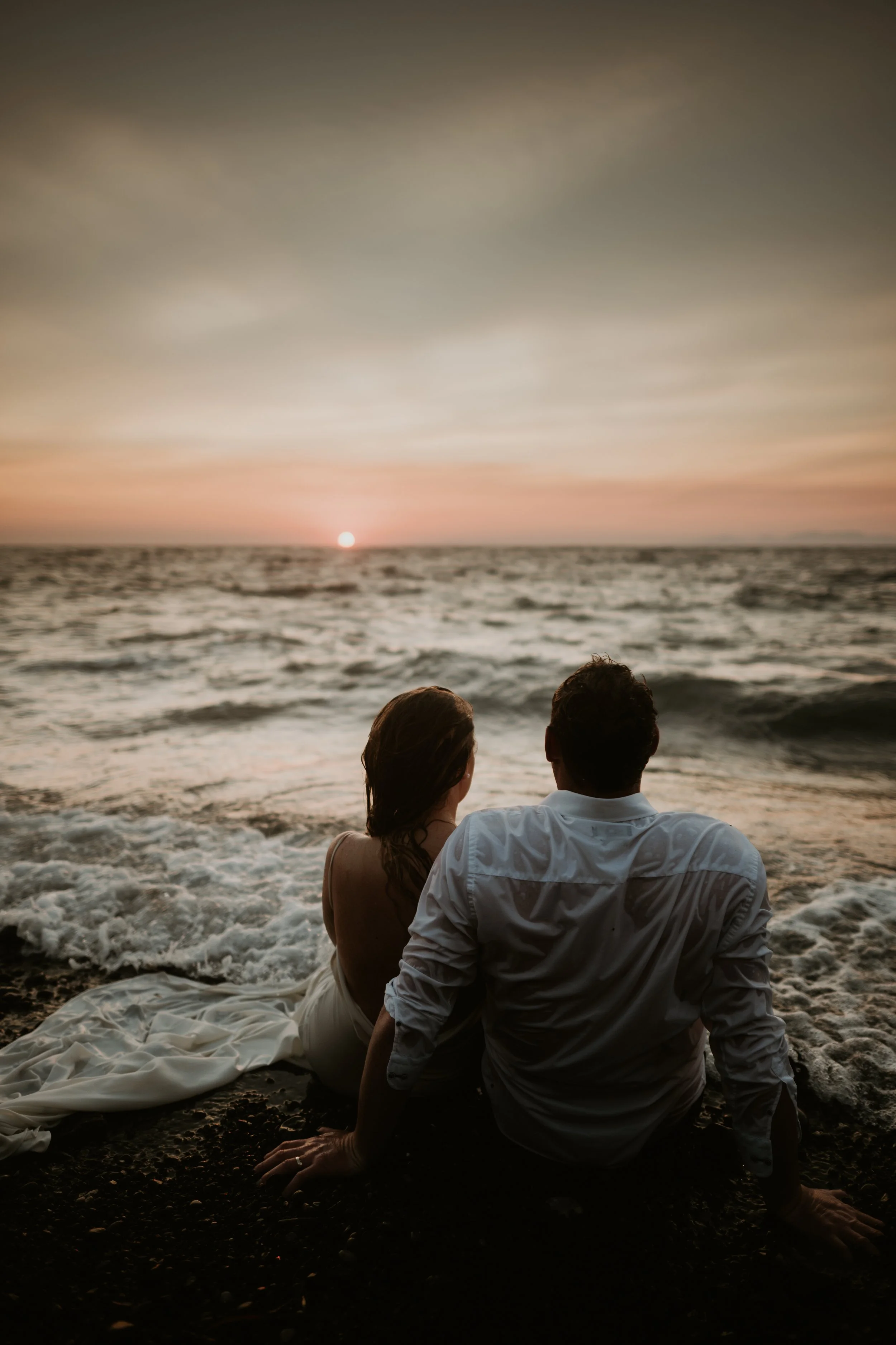 A couple in wedding attire sitting on the beach, watching the sunset over the ocean.