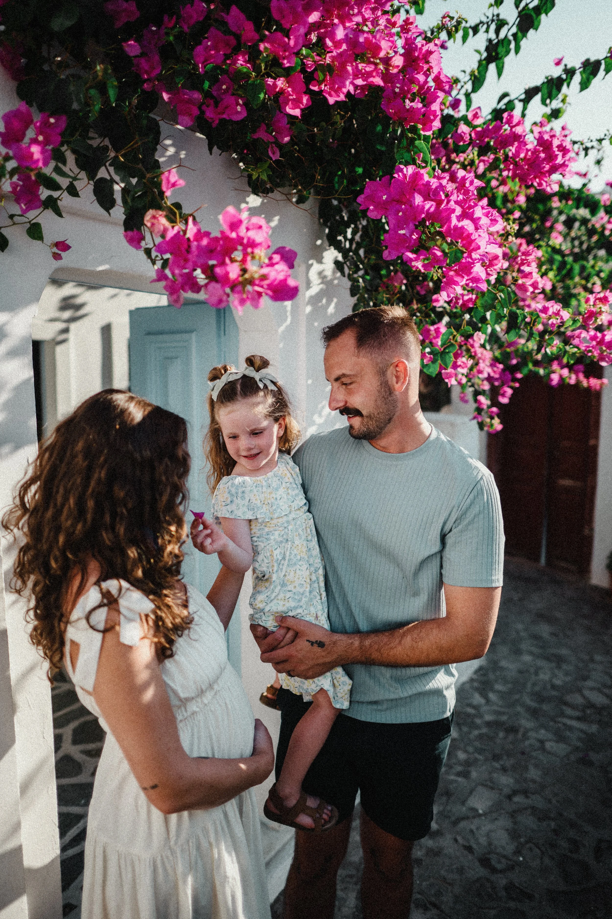 A man holding a young girl, standing outdoors under vibrant pink bougainvillaea flowers, with women nearby and a white building with a blue door in the background.