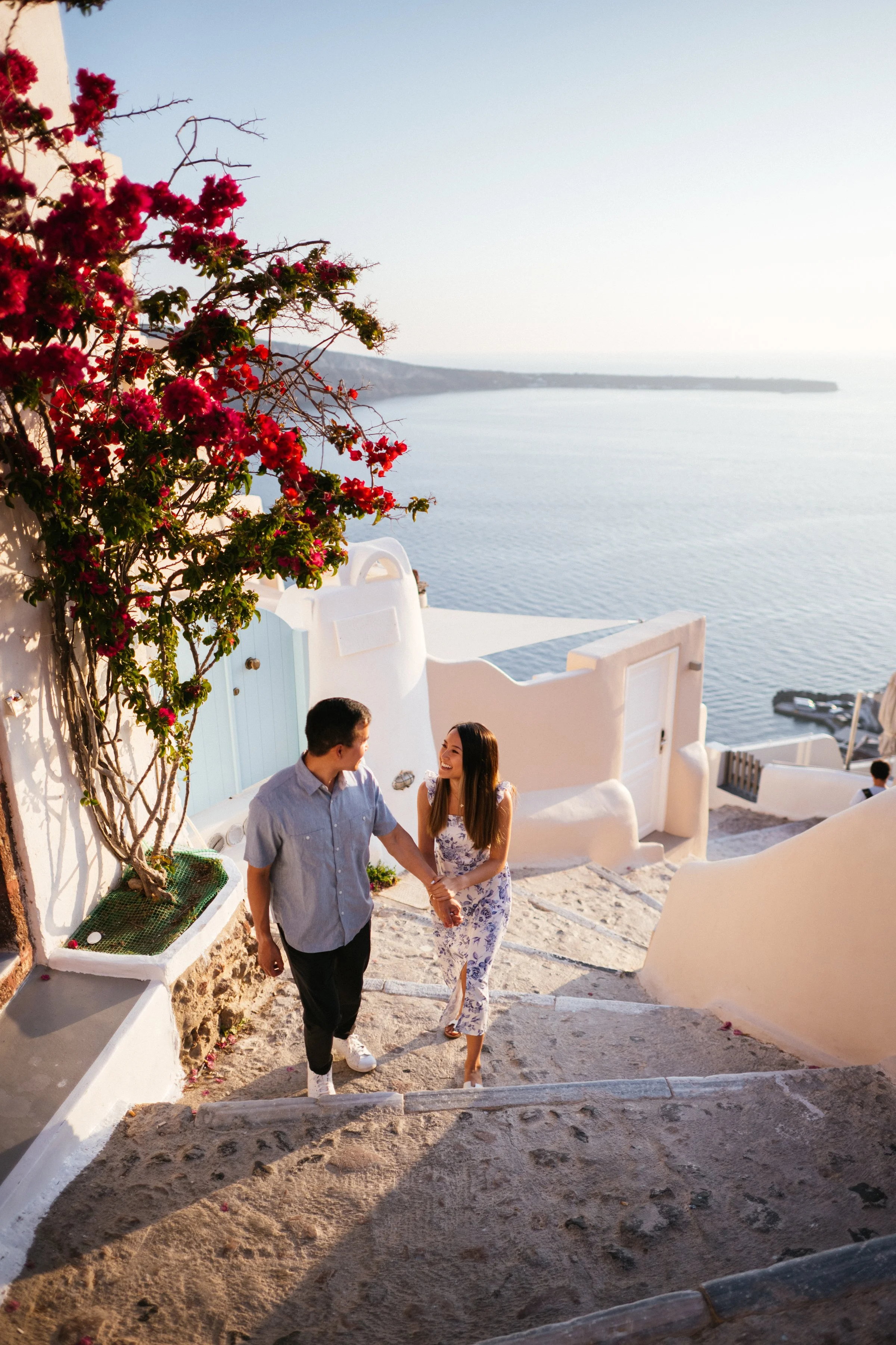 A man and woman holding hands and walking down stone stairs in a seaside location with white architecture and pink flowers, overlooking the ocean during sunset.