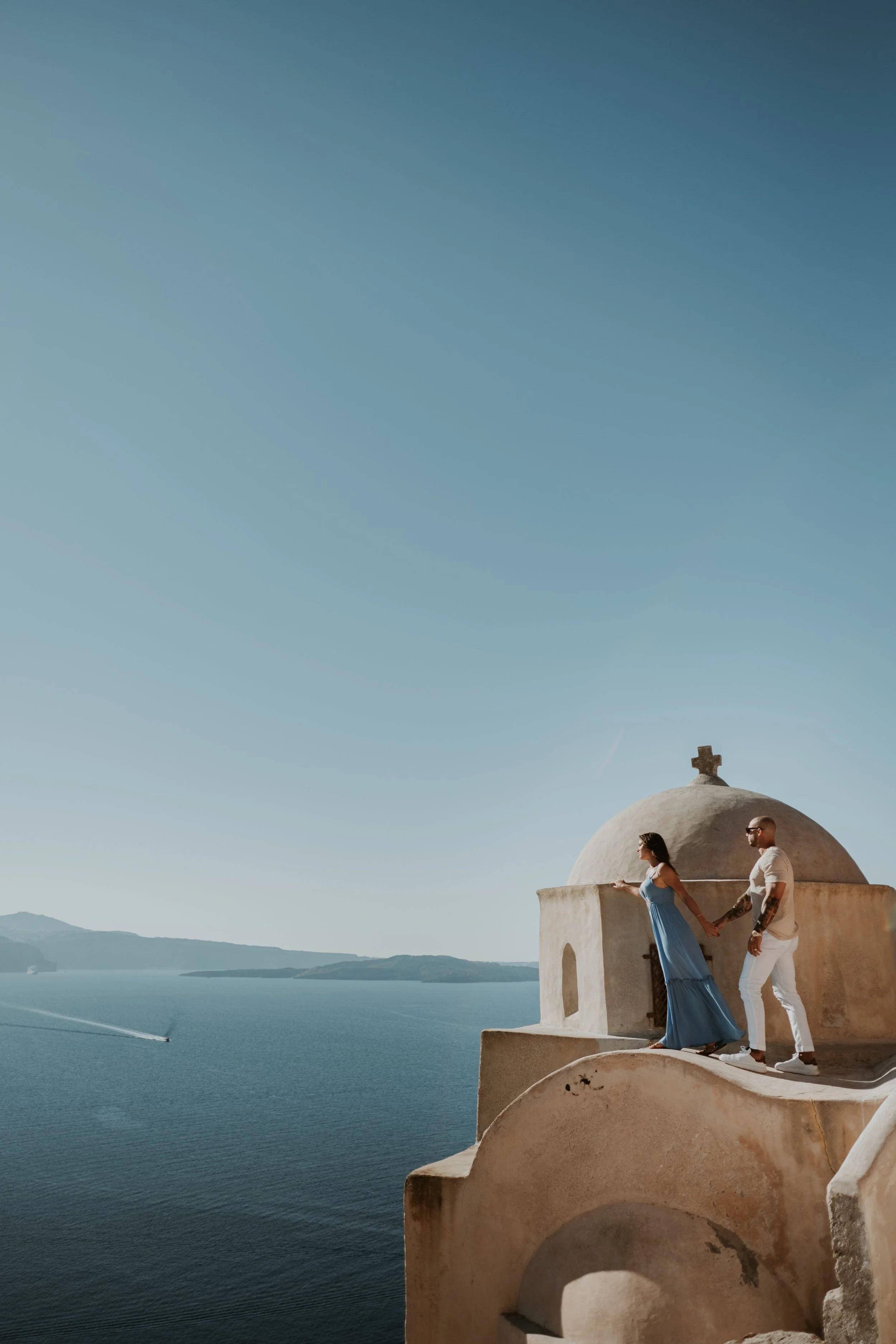 A couple holding hands on a ledge outside a small, white, dome-shaped building with a cross on top, overlooking a body of water and mountains under a clear sky.