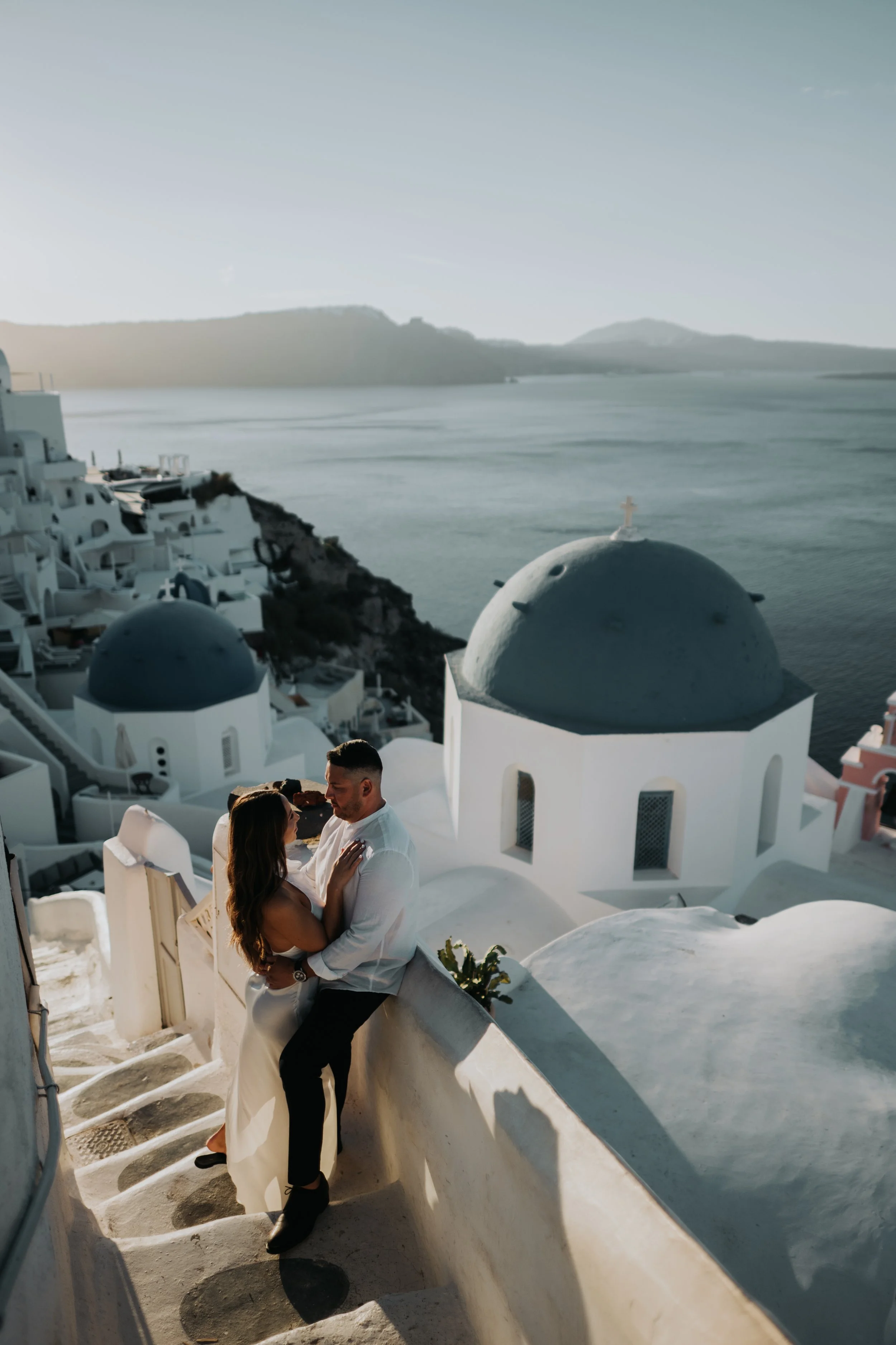 A couple stand close together on white stairs next to a white wall in Santorini, Greece, with a view of the ocean and distant islands in the background, during sunset.