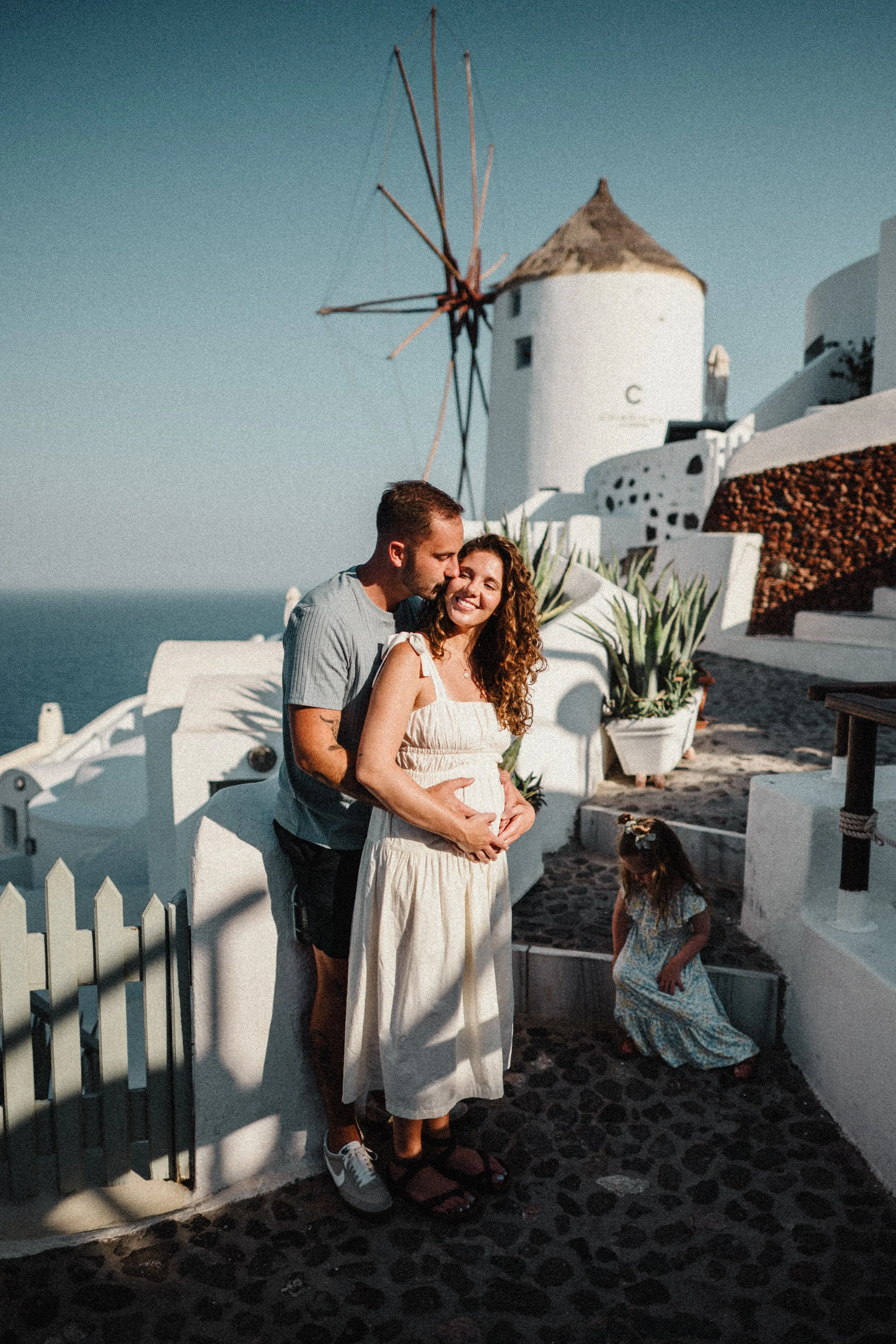 A couple is standing close together on a seaside street, smiling and embracing. The man is kissing the woman's forehead, and she is smiling with her eyes closed and hands on her belly. There is a young girl near them, looking down, dressed in a patte