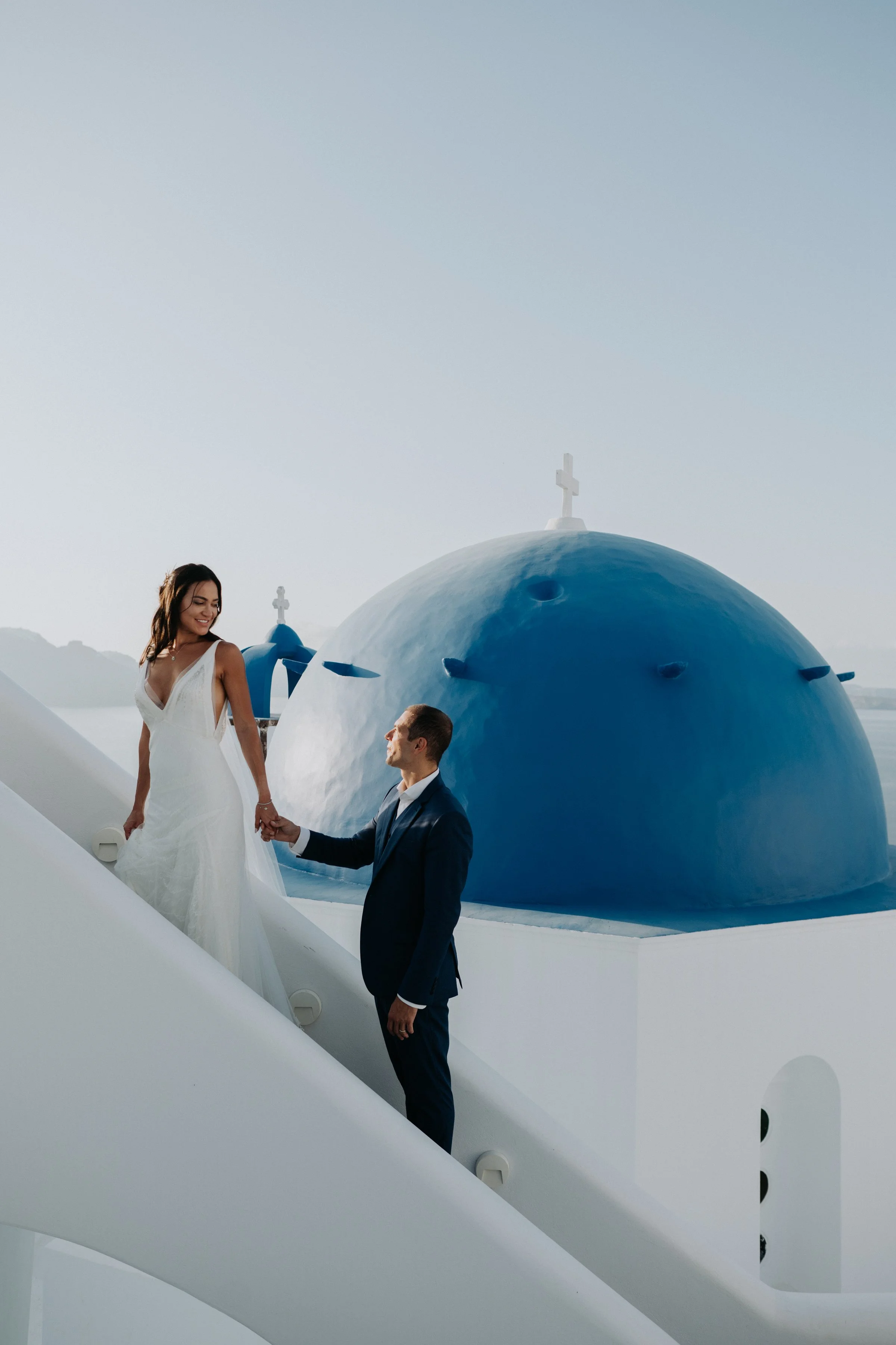 A woman in a white dress and a man in a dark suit, holding hands on a white staircase outside a white building with blue domes and crosses, likely a church, during daylight.