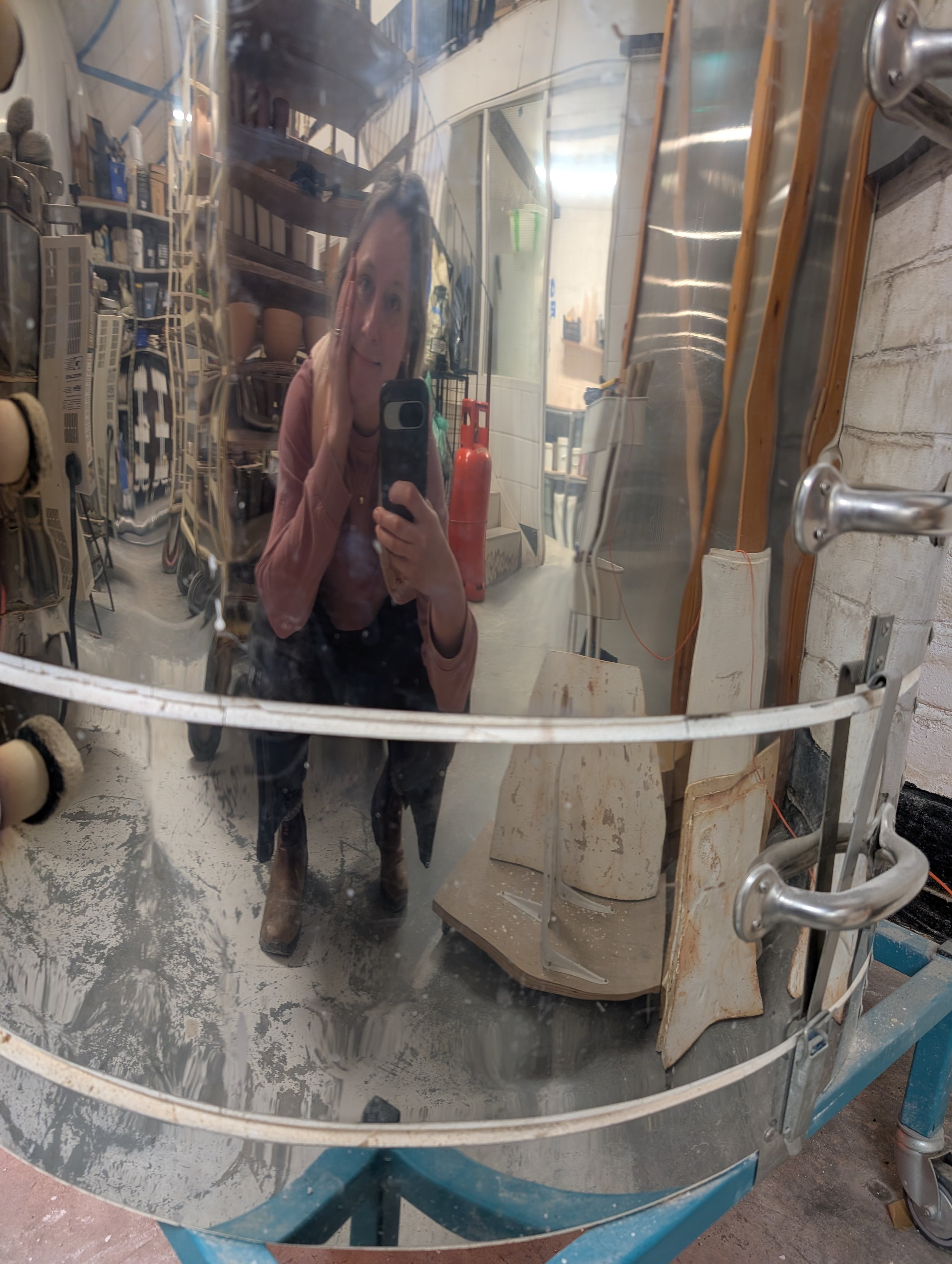 A reflection of a woman taking a selfie with a smartphone, visible in a large, slightly distorted kiln surface in a pottery studio. The background shows shelves, a fire extinguisher, and various tools.