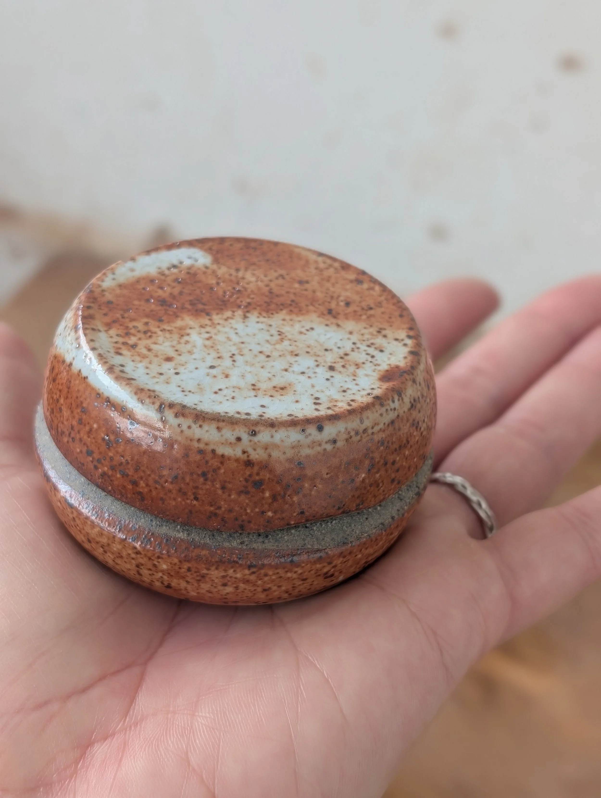Close-up of a hand holding a small, round, brown ceramic mug filled with a frothy beverage topped with cocoa powder or cinnamon.