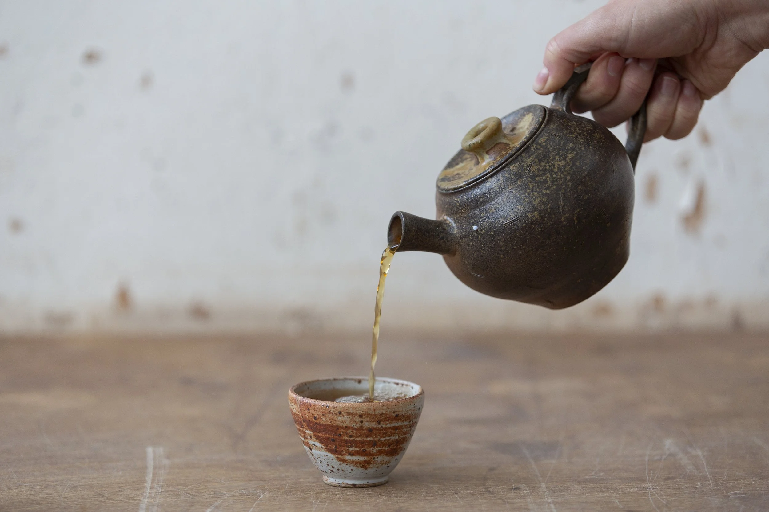 A person pouring hot tea from a rustic, dark ceramic teapot into a small, speckled, red and white teacup on a wooden surface.