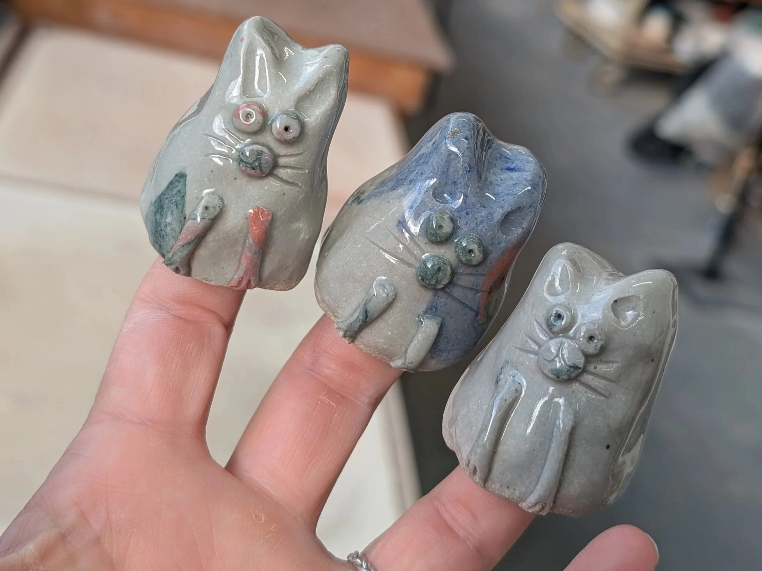 Three ceramic cat figurines in shades of blue and gray, with one showing a front view with pink nose and whiskers, on fingers.