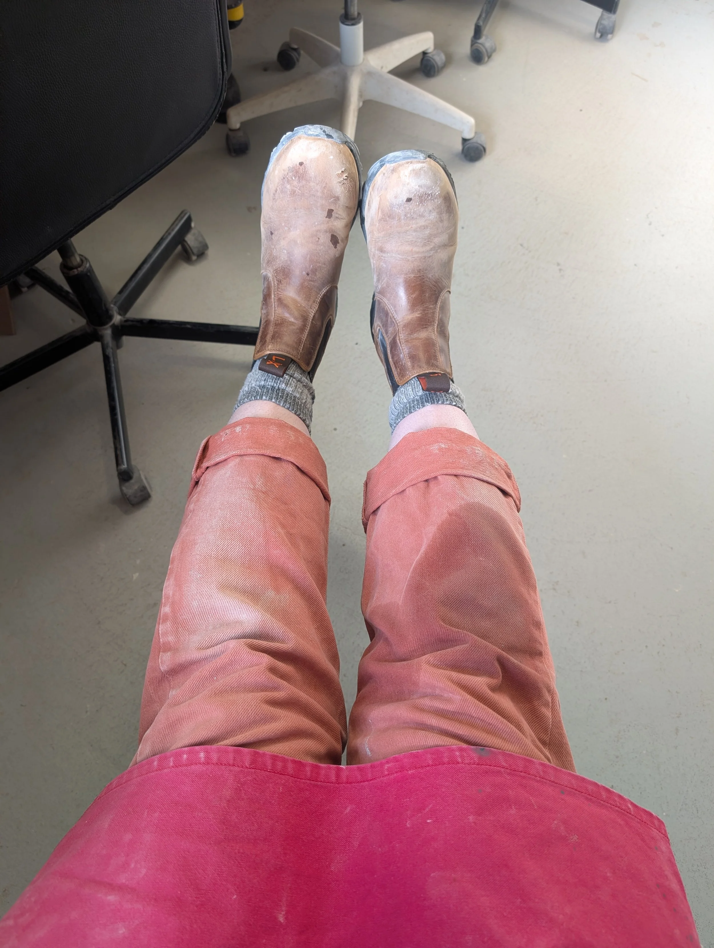 A person wearing brown leather work boots and pink pants sitting with their legs extended, in an indoor workplace setting.