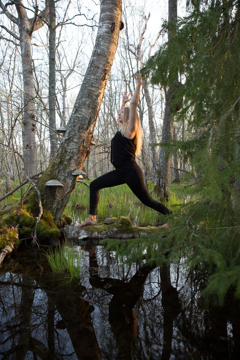A person practicing yoga in a forest, standing in a warrior pose on a mossy log near a pond with trees surrounding them.