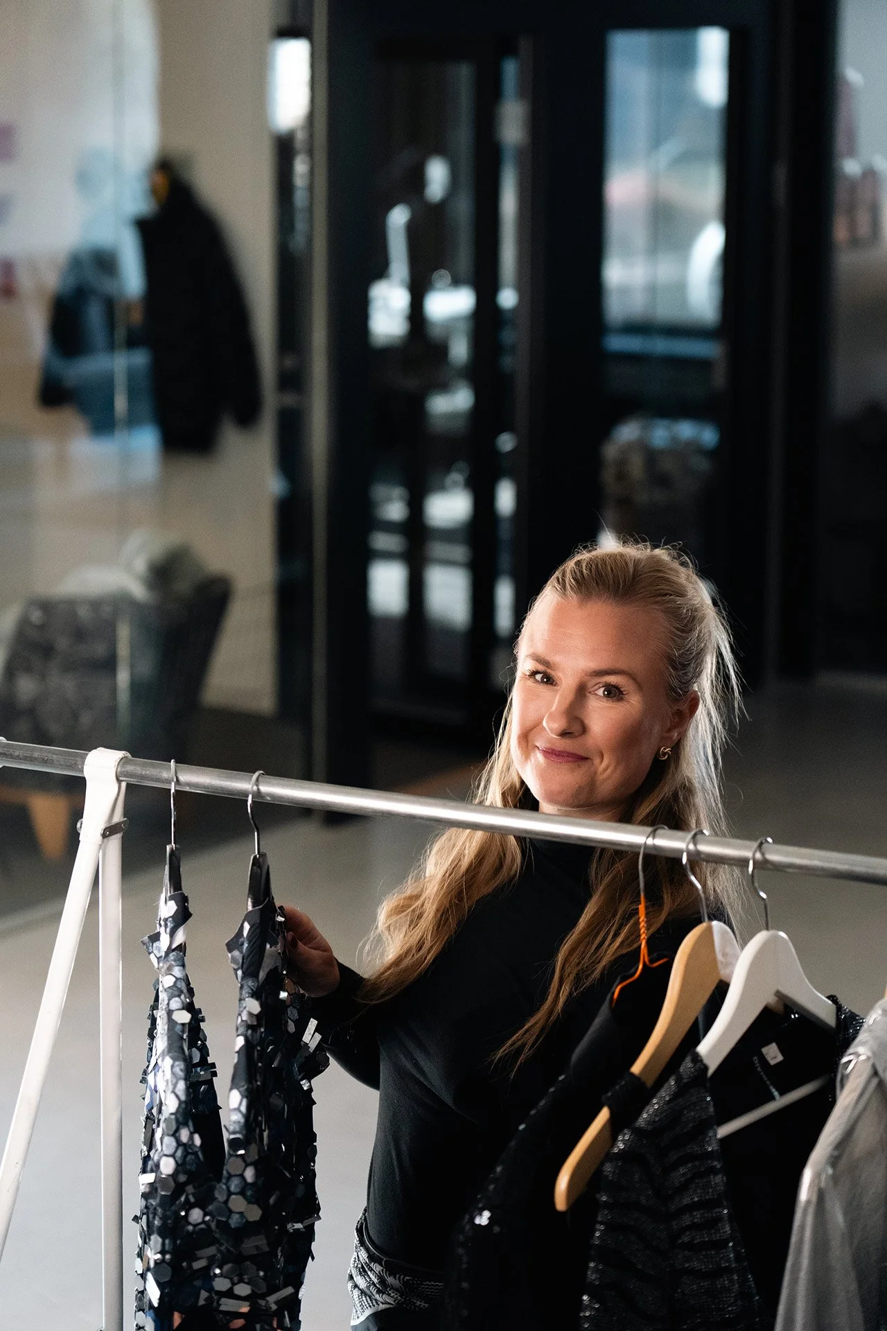 A woman with long, blonde hair smiling at the camera stands behind a clothing rack with various garments, including a black and silver sequined dress. The background features a modern, indoor setting with glass doors and shop windows.