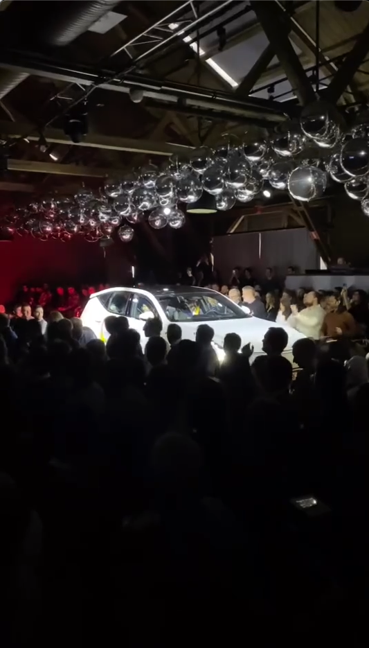 A car standing in a room full of people with the roof decorated with silver balloons.