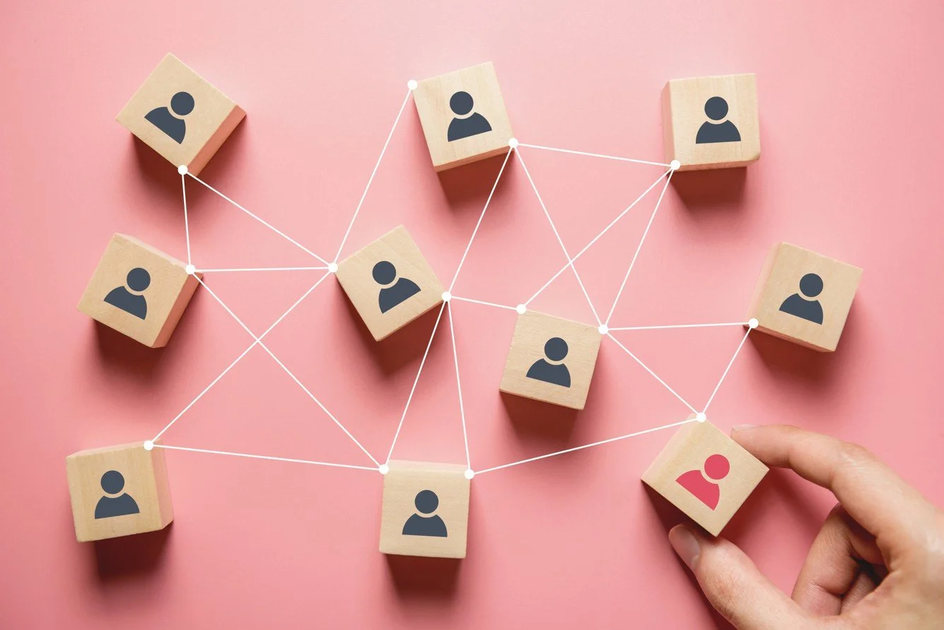 Wooden blocks with user icons connected by white lines, representing social network or connections, on a pink background, with a hand holding one block.