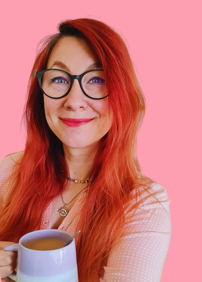 A woman with long red hair and black glasses, smiling, holding a mug of coffee or tea, against a pink background.