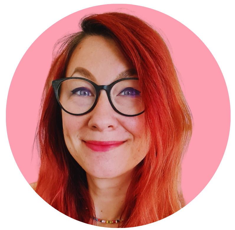 A woman with long red hair, glasses, and a colorful necklace, smiling against a pink background.